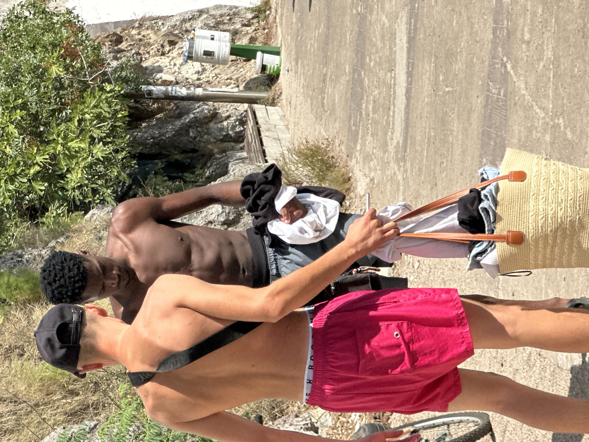 Two young men are walking outdoors on a sunny day in Budva, Montenegro. The man on the left, wearing a black baseball cap and bright pink swim shorts, has a dark strap across his chest. The man on the right, who has dark curly hair and is shirtless, is wearing dark swim shorts. He is holding a bundle of clothes, including a white shirt and a black item, in his right hand. To the right of the men, a large straw bag with brown leather straps is visible on the ground. The bag is filled with clothing. The background shows a rocky cliff face with sparse vegetation and a tree with green leaves. A wooden bench is situated against the rock. A cylindrical trash bin with a white lid and a green base is also visible in the background, as well as a smaller green and white bin. The ground is paved with light-colored cobblestones or concrete. A bicycle wheel is partially visible on the left side of the image.