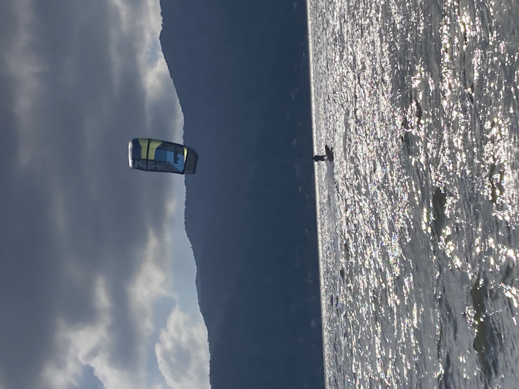 The image depicts a kite surfer on the water against a backdrop of mountains and a cloudy sky. The location is likely in Florianópolis, Brazil, given the landscape. The water is reflective, suggesting sunlight. A kite is in the air.