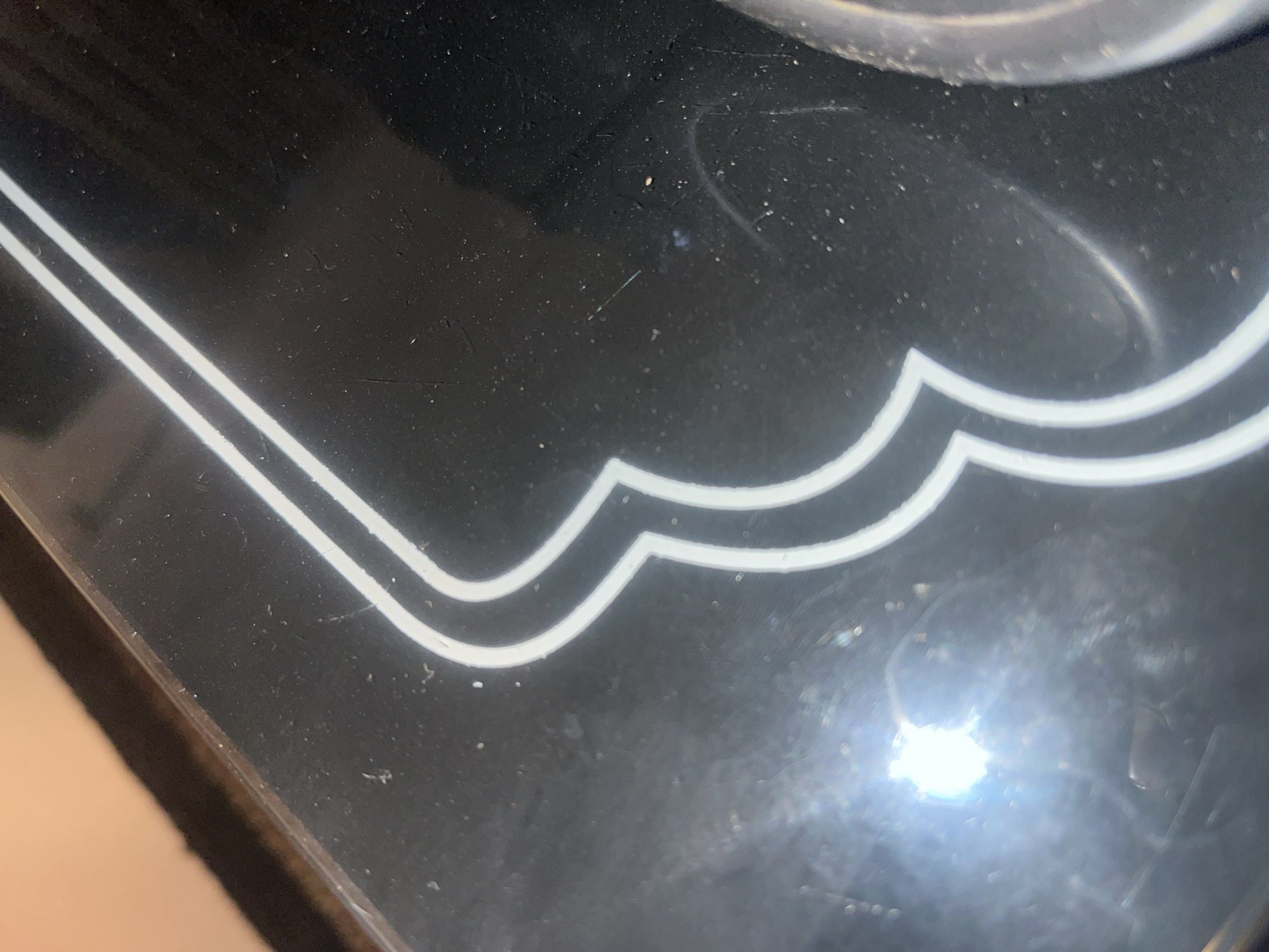 The image shows a close-up, oblique view of a shiny black surface with a decorative white line pattern. The pattern consists of two parallel, curved white lines forming a stylized scalloped or cloud-like shape. The surface appears to be dusty or smudged, with small white specks visible throughout. There is a bright, blown-out light source in the lower left quadrant, creating a strong glare and obscuring details in that area. A circular, metallic or clear plastic edge is visible on the far right side, suggesting the surface may be part of a manufactured object like a tabletop, appliance, or decorative panel. The background, visible in the upper left, is out of focus and appears to be a warm, light brown color, possibly a textile or wall. The overall impression is of a detailed view of a surface that is in need of cleaning, with elements of its design and texture highlighted by artificial light. The image does not contain any identifiable landmarks or context that would allow for its specific location within Jalingo, Nigeria.