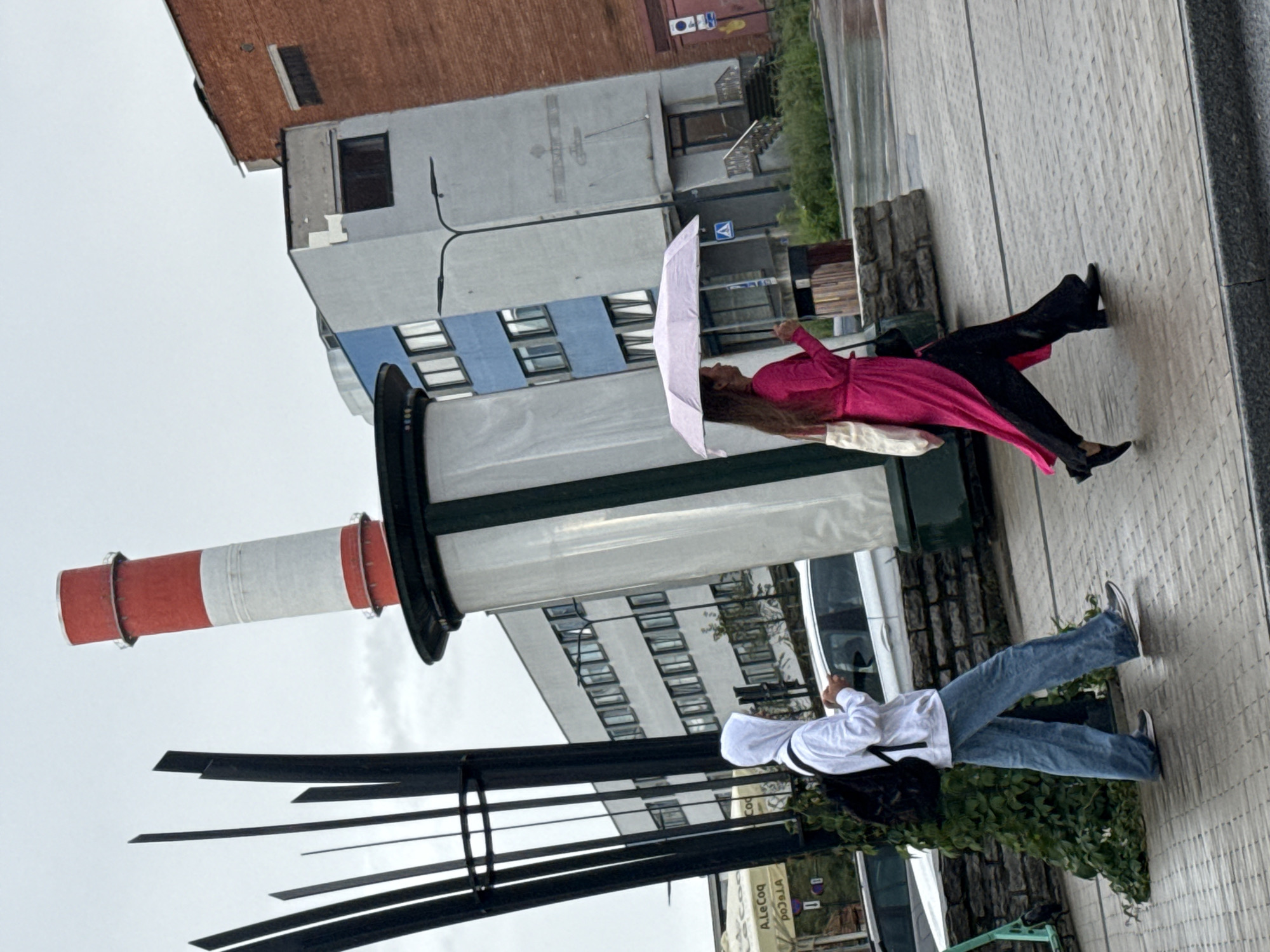 An overcast day in Tallinn, Estonia, features two individuals walking on a wet pavement. In the foreground, a person in a white hooded sweatshirt and loose-fitting blue jeans, carrying a black backpack, is looking down at their phone. To their right, a person in a bright pink dress and black pants walks under a light pink umbrella, carrying a cream-colored tote bag and a black purse. In the background, a towering cylindrical structure with red and white bands is prominent. Behind it, a building with blue and grey sections stands next to a red brick building. A metal sculpture with several vertical black elements is situated to the left of the cylindrical structure. Cars are visible parked in the background, and the pavement appears damp, suggesting recent rain.