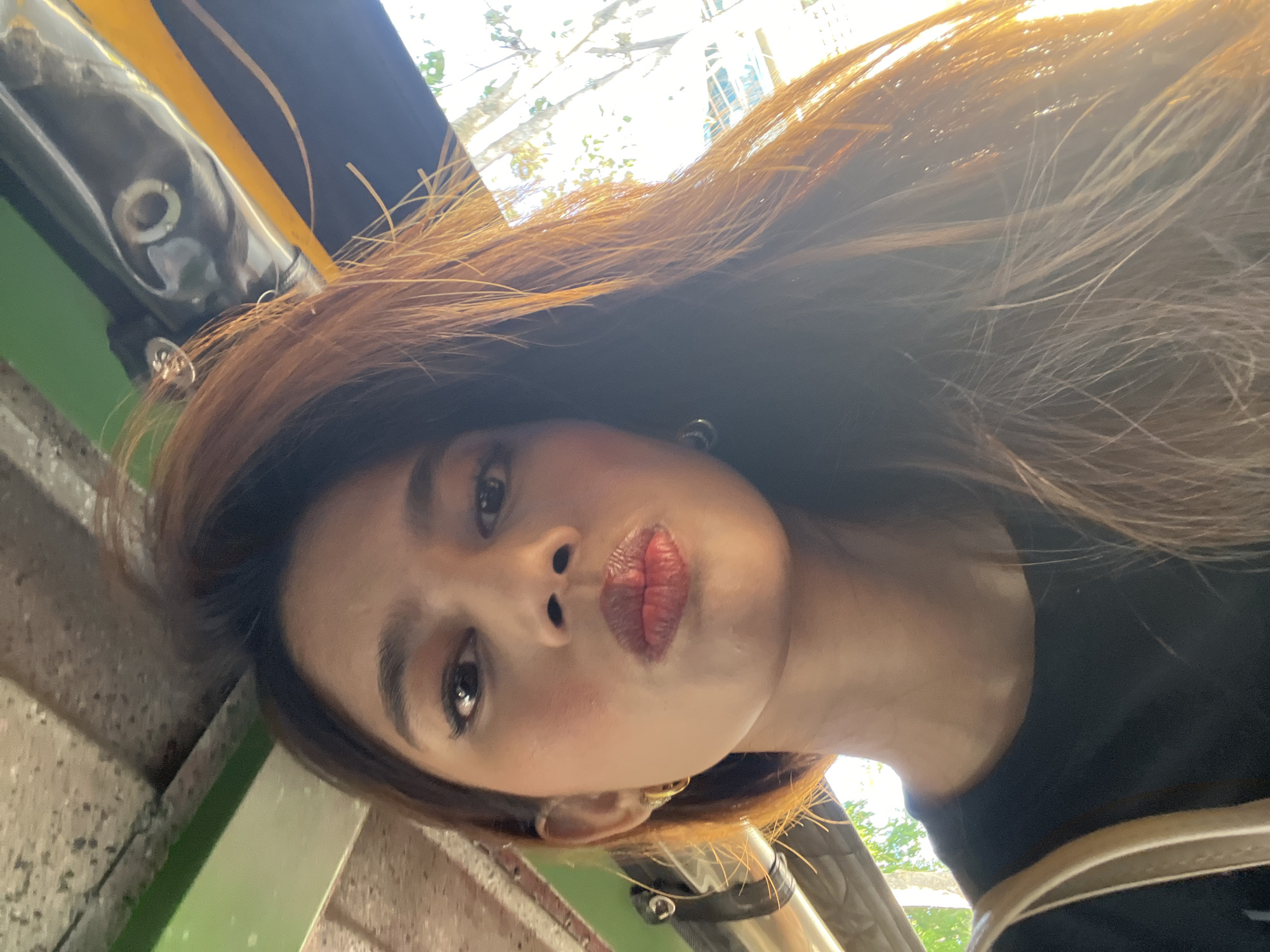 The image is a selfie taken from inside a vehicle, likely a tricycle common in Davao City, Philippines, given the overhead structure visible. The subject is a woman with brown hair, applying lipstick. She is wearing a black t-shirt and gold hoop earrings. The background shows a glimpse of the outdoors, with trees and sunlight filtering through. The overall lighting suggests it's daytime.