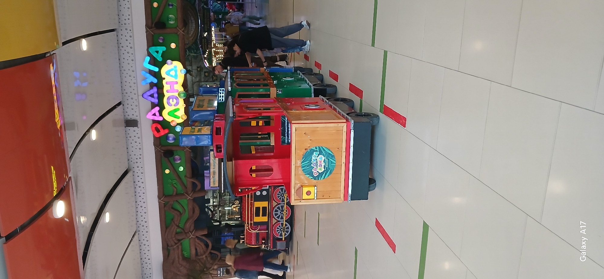 This photograph shows a colorful children's train ride in a shopping mall in Yekaterinburg, Russia. The train is composed of multiple cars in red, green, blue, and wood colors, each decorated with various designs. It is placed on a white tiled floor marked with green and red lines. The background features a decorative wall with a green tree-like structure and illuminated lettering in Cyrillic script, spelling out the name of the attraction. Several people are present, including a couple in casual clothing, possibly overseeing or waiting for children to use the train ride. The lighting is bright, indicative of an indoor commercial space. A 'Galaxy A17' watermark is visible in the lower right corner.