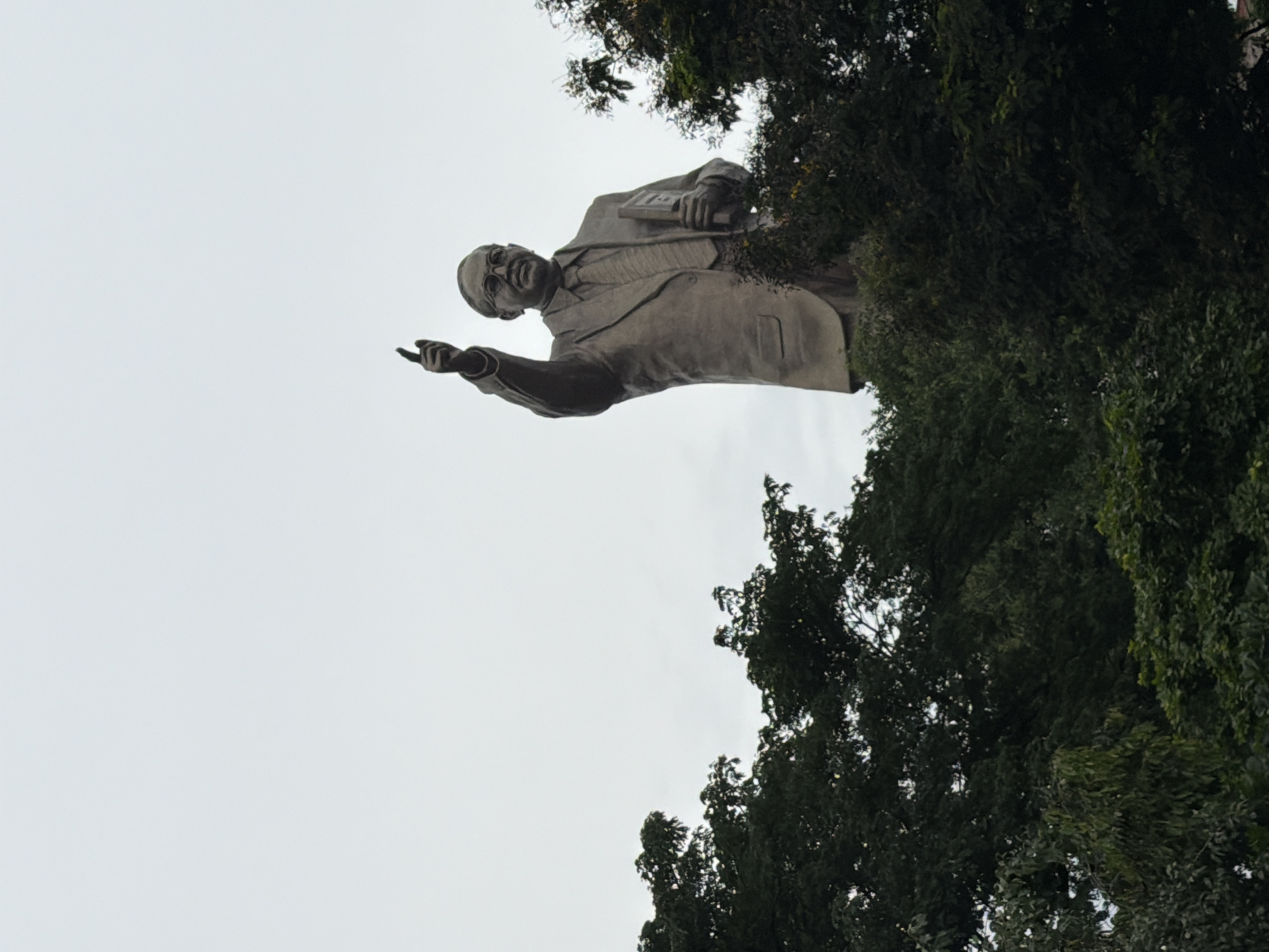The image shows a statue of Dr. B.R. Ambedkar, a prominent Indian jurist, economist, politician, and social reformer, situated in the city of Hyderabad, India. The statue is made of bronze and depicts Ambedkar in a suit, holding a book in his left hand and pointing upwards with his right index finger. He is wearing glasses. The statue is positioned against a pale, overcast sky, and is partially obscured by lush green trees and foliage in the foreground and on the sides. The composition suggests that the statue is located in a public space, possibly a park or a memorial, given the presence of trees. The lighting is diffused, indicating an ambient daylight condition.