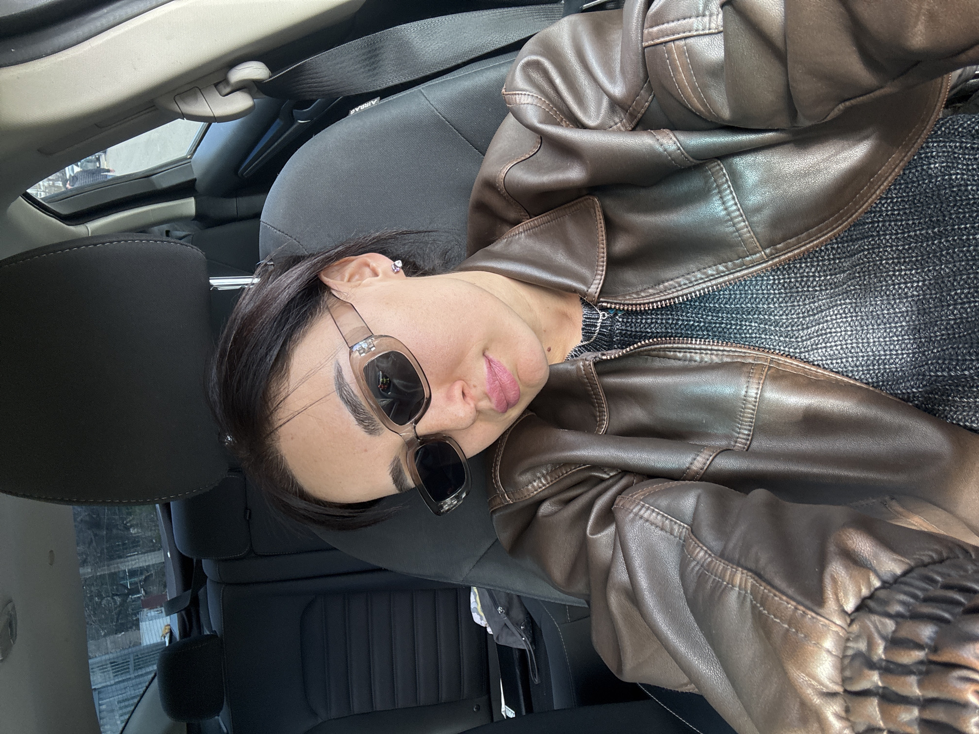 A person is seated inside a car, likely in Tbilisi, Georgia, judging by the exterior view from the window. The individual is wearing sunglasses and a brown leather jacket over a gray sweater. The interior of the car is black, with visible headrests, a safety belt, and parts of the car's ceiling and door panel. The outside scene through the car window shows buildings and trees. The person appears to be taking a selfie or photo inside the vehicle.