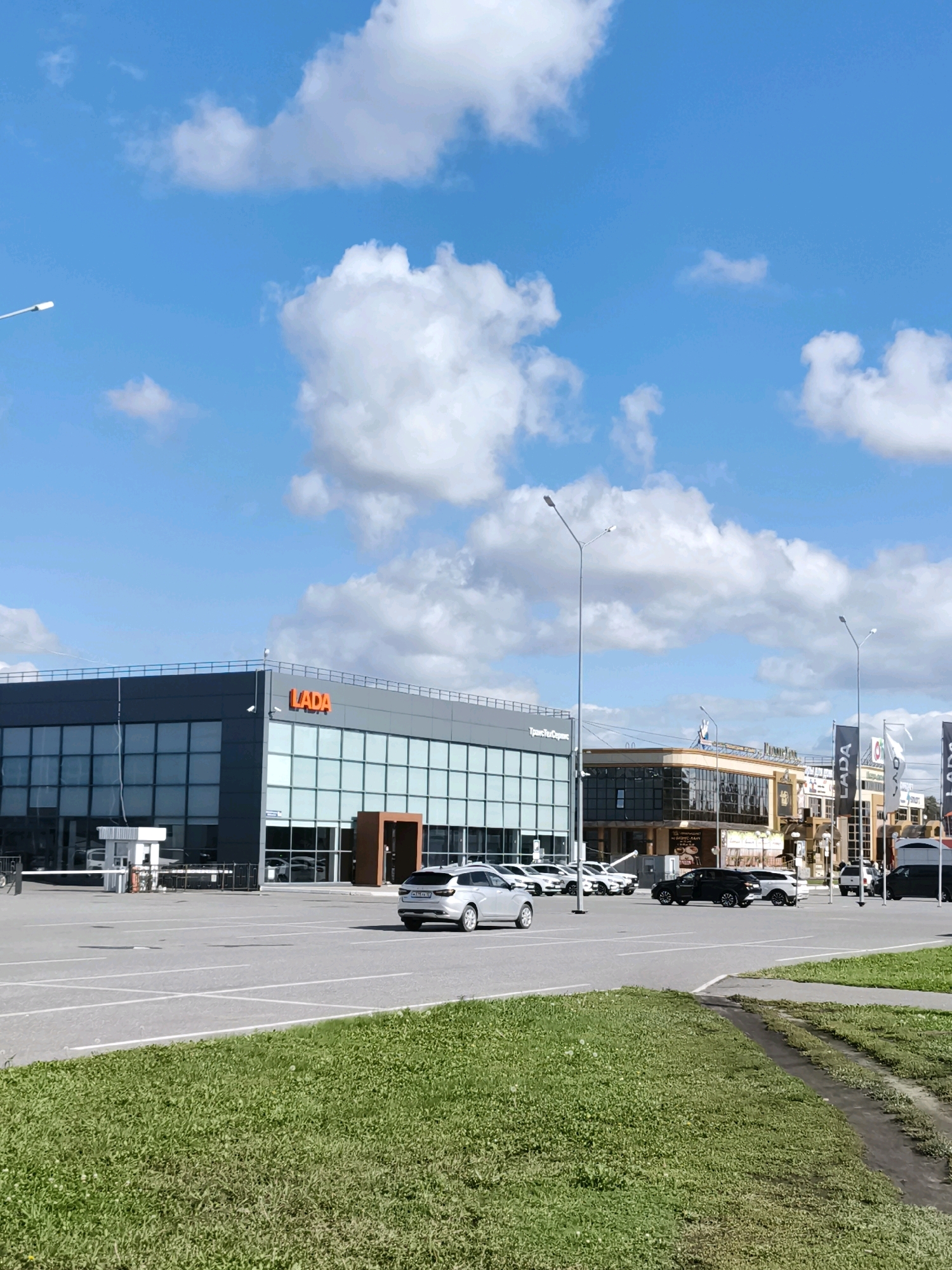 The image shows a LADA car dealership in Yoshkar-Ola, Russia, on a clear day with a blue sky and white clouds. The dealership building has a modern design with large glass windows and the LADA logo prominently displayed. In front of the dealership, there is a parking lot with several cars, including LADA models. Some cars are parked, while others appear to be in the process of being driven. A grassy area with a dirt path is in the foreground, suggesting a natural element within the urban landscape. The overall scene depicts a commercial area with a focus on automotive sales.