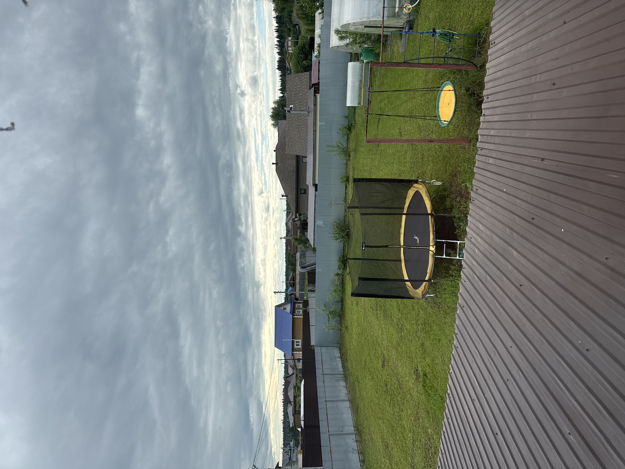 The image depicts a residential backyard scene in Сизёво, Russia, captured during daytime under an overcast sky. The viewpoint is elevated, looking down over a brown corrugated metal roof that occupies the bottom portion of the frame. The roof is secured with visible fasteners.
Beyond the roof, a well-maintained green lawn fills the midground, enclosed by a corrugated metal fence of varying shades of grey-brown and light blue. Within the lawn area, a black and yellow trampoline with a safety net and a white two-step ladder is positioned on the left. To its right, a red-framed swing set holds a single yellow disc swing suspended by black ropes. Further to the right, a blue metal ladder with a green hose coiled around it and a green barrel are visible near the fence. Two tunnel-shaped greenhouses with translucent coverings are situated along the right side of the fence line.
In the background, beyond the immediate yard and fences, various residential houses are visible under the cloudy sky. Notable structures include a wooden house with a bright blue metal roof on the left side of the frame and a larger house with a brown shingle roof dominating the central background. Utility poles and power lines are present on the left. A dense line of trees and distant forest can be seen on the horizon, indicating a rural or semi-rural setting. No individuals or active interactions are present in the scene. The lighting is diffuse, consistent with heavy cloud cover.