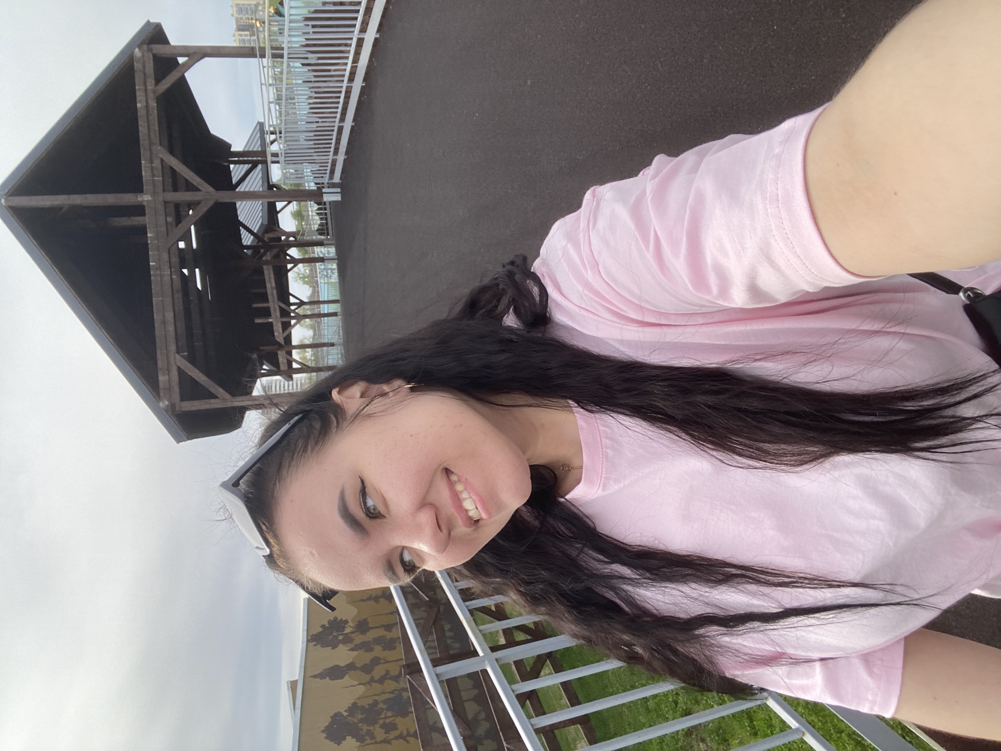 A young woman takes a selfie outdoors. She is wearing a pink t-shirt and sunglasses perched on her head. She has long, dark, wavy hair. In the background, there is a wooden structure resembling a gazebo or shelter, a metal railing, and some greenery. The location is in Perm, Russia, as evidenced by the mural depicting a forest scene in the lower left. The scene appears to be near a river or lake, given the glimpses of water visible in the distance through the wooden structure. The weather seems overcast.