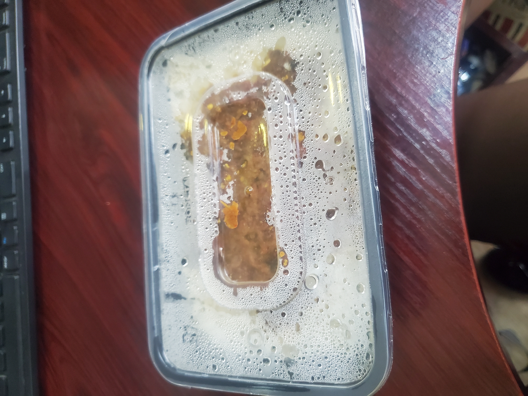 A clear plastic food container is prominently featured, centered on a reddish-brown wooden surface, likely a table or desk. The transparent lid of the container is covered with numerous small droplets of condensation, suggesting the contents are warm.
Inside the container, a portion of white food, consistent with rice, fills the main compartment. A raised, rectangular section within the lid contains a darker, brownish-orange, chunky substance, which appears to be a sauce or stew with visible flecks of ingredients. This smaller compartment is designed to keep the sauce separate from the main food item.
To the left of the container, the edge of a black keyboard is partially visible. On the right side, an out-of-focus, dark-colored object, possibly a person's leg or arm, or another item, is seen in the background. The wooden surface displays a diagonal grain pattern.
There is no discernable text on the food container or the visible part of the keyboard due to reflections and resolution. The lighting indicates an indoor setting. The image captures a ready-to-eat meal, potentially a takeaway or meal prep, placed on a surface in what could be a home or office environment in Gwagwada, Nigeria.