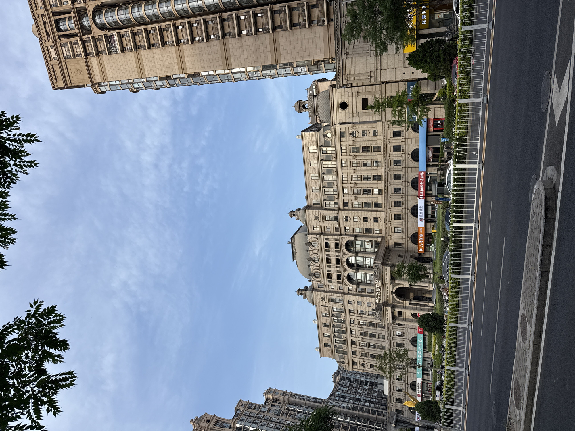 The image captures a street scene in Chaoyang District, China, showcasing buildings and street elements. The shot is taken from a low angle, emphasizing the height of the buildings. One building displays a facade of numerous windows and architectural details, and other building is modern-looking and also tall. A street runs alongside the buildings, delineated by a white metal fence. Some greenery is visible, including trees, and the blue sky. Chinese text is present on some of the buildings.