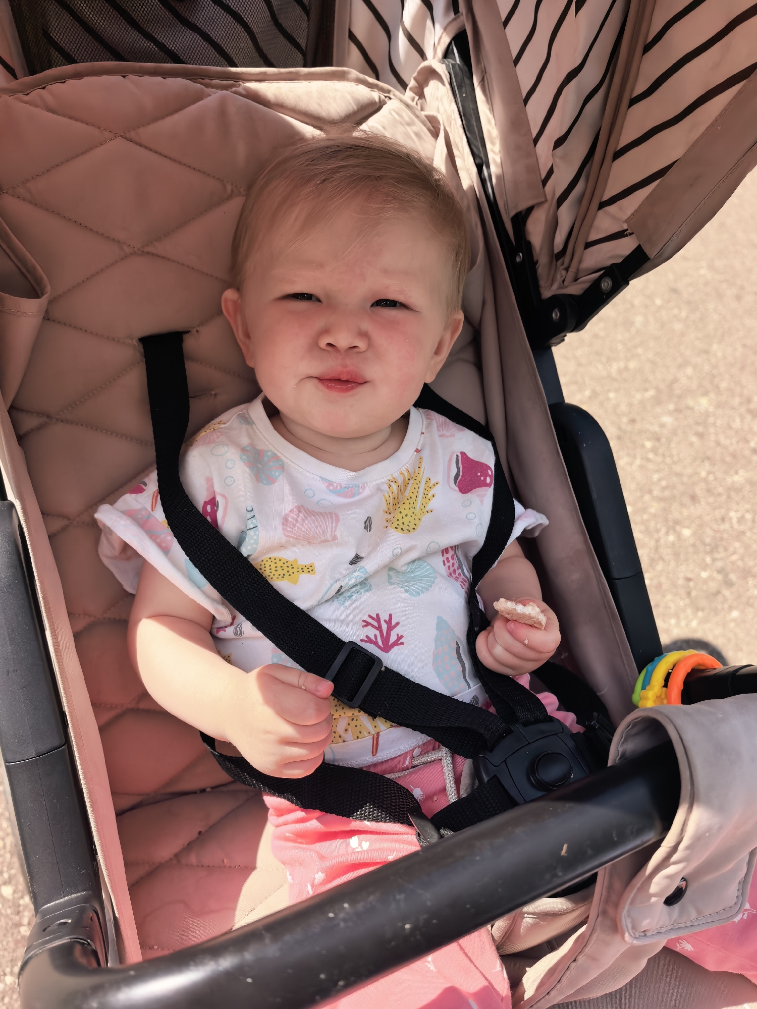 A young child is seated in a stroller. The child has blonde hair and is wearing a white t-shirt with a seashell pattern and pink shorts. The child is holding a piece of food in their right hand. The stroller has a pink quilted interior and a striped canopy. A multi-colored toy is attached to the stroller's handle. The background appears to be an outdoor environment with a sandy or gravelly surface. This image does not contain any information to determine its location in Sterlitamak, Russia.