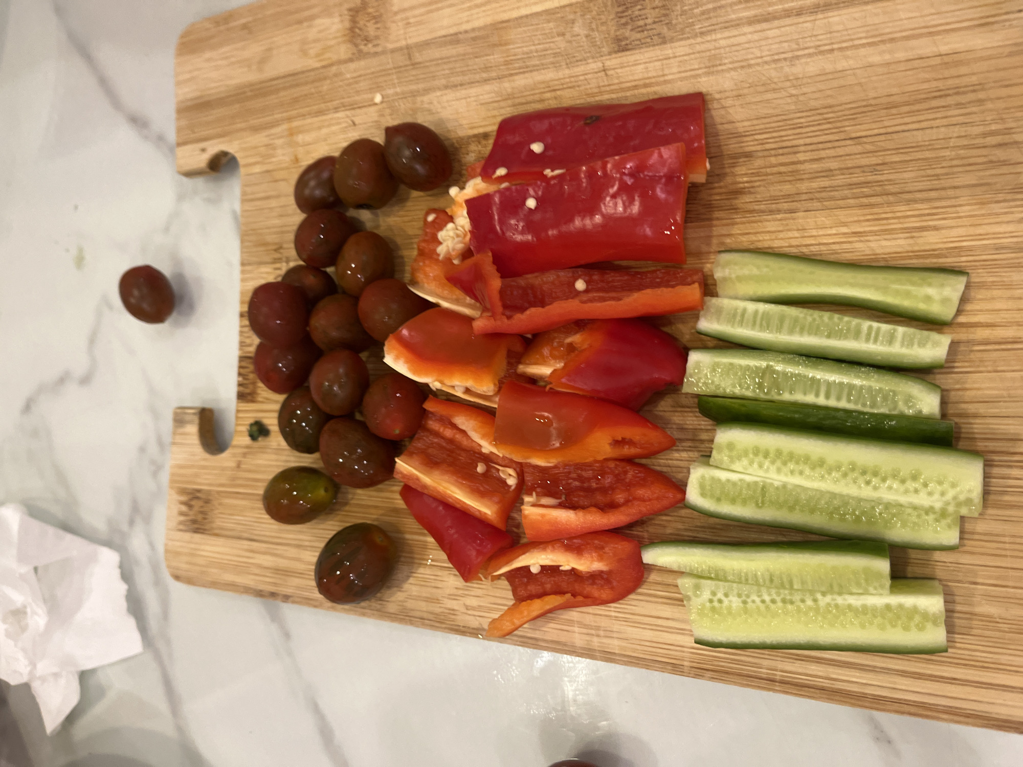 The image shows a wooden cutting board with an assortment of vegetables arranged on it. There are dark-colored cherry tomatoes, sliced red bell peppers, and cucumber sticks. There is a crumpled white paper towel adjacent to the cutting board. There are no people present in the image.
There is no information to suggest this image was taken in Bac Nha Trang Ward, Vietnam.