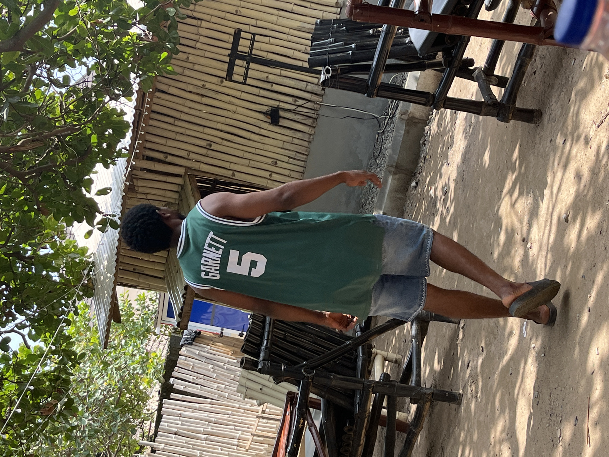 The image depicts an outdoor scene, likely in Abuja, Nigeria, where a young Black male wearing a green basketball jersey with "GARNETT 5" printed on the back and denim shorts is walking away from the camera. He is wearing dark slippers. The background features a structure made of bamboo, with a corrugated metal roof, and a chain-link fence. There are also several pieces of black metal furniture, possibly chairs, in the foreground and midground. Green foliage from a tree hangs overhead, casting shadows on the sandy ground. The environment appears rustic and possibly a casual outdoor gathering spot.
