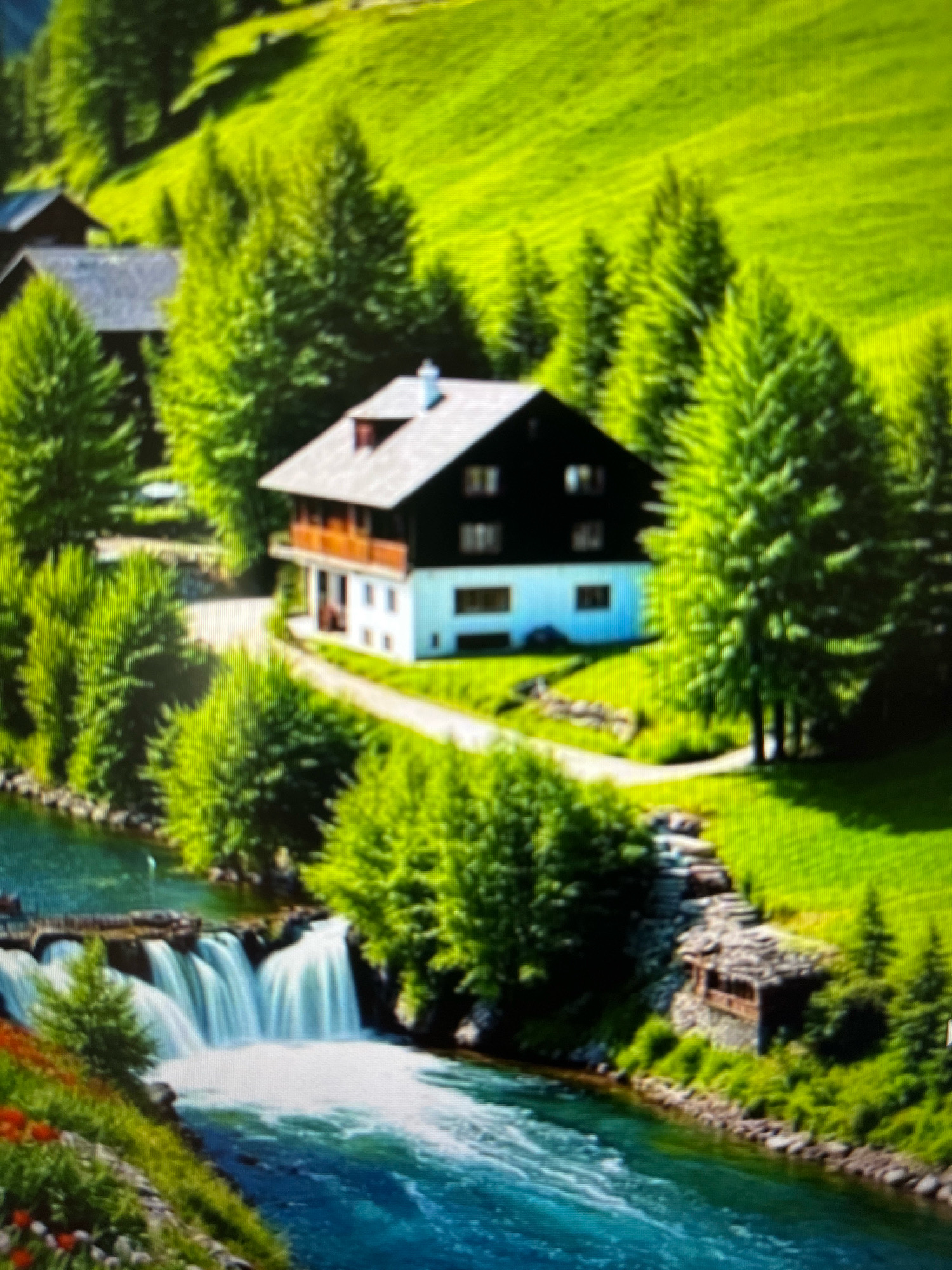 The image depicts a picturesque landscape in a mountainous region, featuring a multi-story house with dark siding and a white lower level, surrounded by lush green trees and rolling hills. A river with a waterfall flows in the foreground, bordered by rocky terrain and vegetation. There are no people visible in the image, nor any actions or interactions. The scene appears to be a serene, natural environment. The location is not in Novocheboksarsk, Russia.
