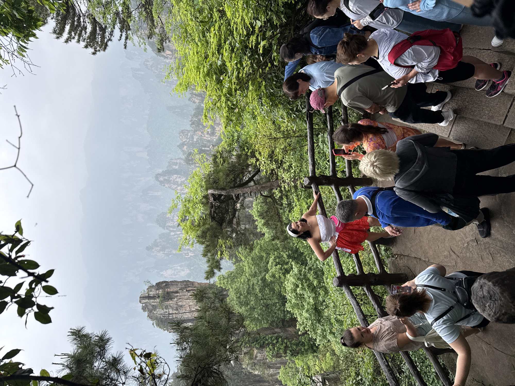 The image depicts a group of tourists viewing a scenic overlook in Wulingyuan, China. A woman in a red skirt and white top poses on a wooden railing. Several other tourists, visible on the path, are observing the scenery, taking photos, or talking. The background features the characteristic sandstone pillars and lush vegetation of the Wulingyuan area. The atmosphere suggests a somewhat overcast or misty day.