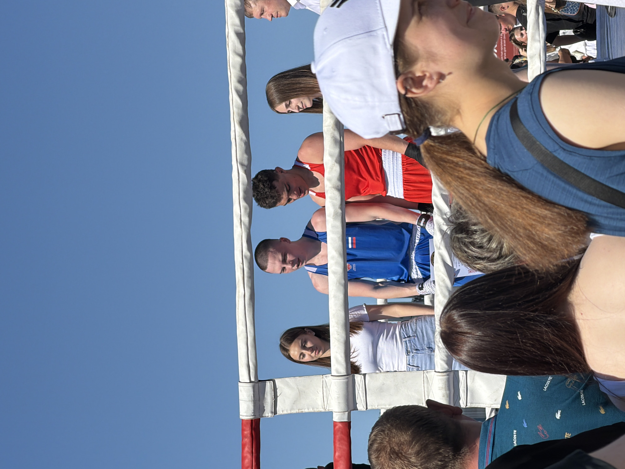 The image depicts a boxing ring viewed from an audience perspective. Two boxers are visible in the ring, one wearing a red uniform and the other a blue one with the Russian flag. Several spectators are standing around the ring, observing the action. The setting appears to be outdoors under a clear blue sky. The specific location is Samara, Russia, inferred from the Russian flag on the blue uniform. The boxers are potentially interacting, engaging in a boxing match, while the spectators are observing the activity.