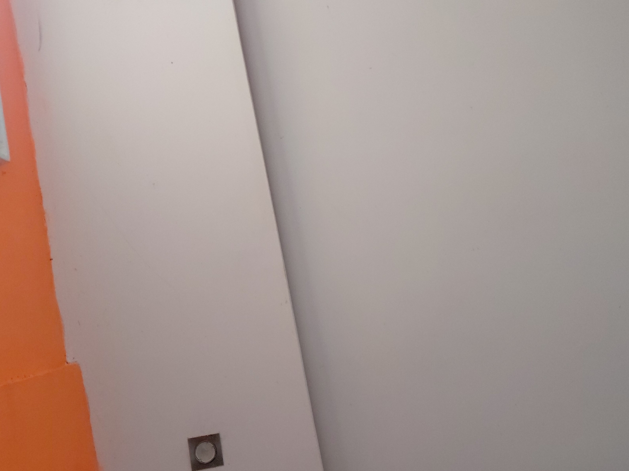 The image displays an interior scene, likely a room or hallway in Santa Ana, Venezuela, characterized by its distinct wall coloring and architectural lines. No people are visible.
The left side of the frame features a wall painted in a vibrant, uniform orange. This orange section is sharply delineated, creating a clean vertical edge where it meets a much lighter, off-white or light grey wall that dominates the rest of the image.
Running diagonally from the top center to the bottom right, there is a pronounced seam, crease, or the edge of another architectural element, possibly indicating a corner or a panel division. This diagonal line separates two slightly different shades or planes of the light grey/off-white surface, with the area to its right appearing marginally darker or in shadow.
Near the bottom-left corner of the lighter wall, close to the orange section, a small, square-shaped fixture is visible. It features a metallic or silver-colored frame with a circular, slightly reflective element in its center, possibly an electrical outlet, switch, or a small recessed light.
The overall impression is one of a modern or simply designed space, with the contrasting colors providing a visual dynamic. There is no specific activity taking place, and no visible text in the image. The lighting appears even, suggesting indoor artificial light or diffuse natural light.