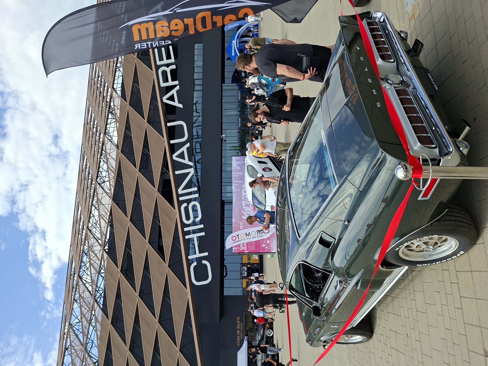 The image shows an outdoor event at the Chisinau Arena, located in the city of Stăuceni, Moldova. In the foreground, a dark green classic car, likely a Ford Mustang Shelby, is parked and cordoned off with a red ribbon. The car's rear features prominent "SHELBY" badging and distinctive taillights. Several people are visible in the background, some looking at cars and others standing around. The Chisinau Arena building serves as the backdrop, with its distinctive geometric patterned roof and large white lettering spelling out "CHISINAU ARENA." A tall black flag with orange lettering is also visible on the right side of the image. The ground is paved with interlocking bricks. The sky is partly cloudy.