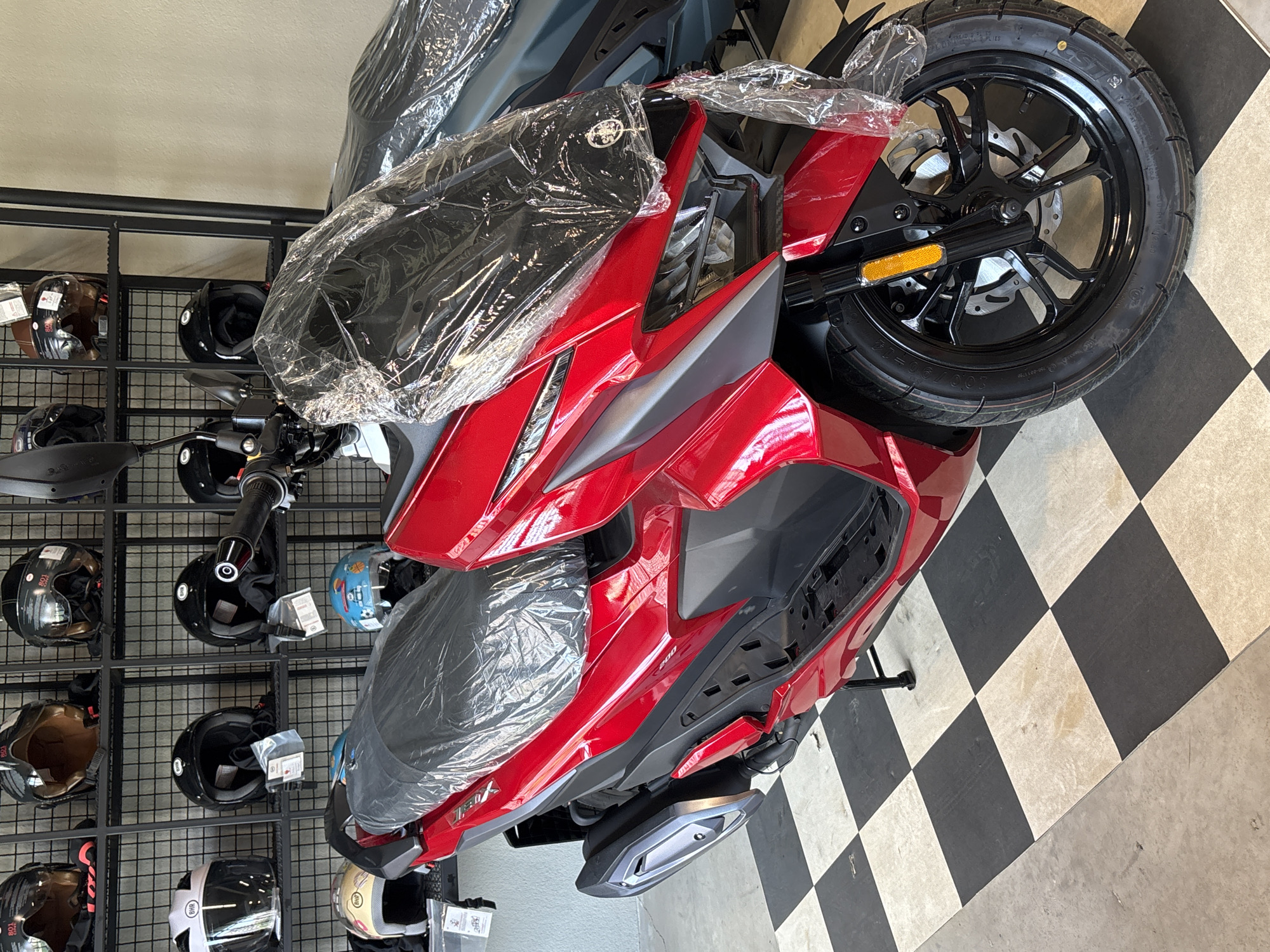 The image shows a red scooter parked in what appears to be a showroom or retail space. The scooter is covered in clear plastic. Behind the scooter, shelves are visible, stocked with various motorcycle helmets. The floor is a black and white checkered pattern. The scene is well-lit and the scooter is the primary focus. No people are visible in the image.