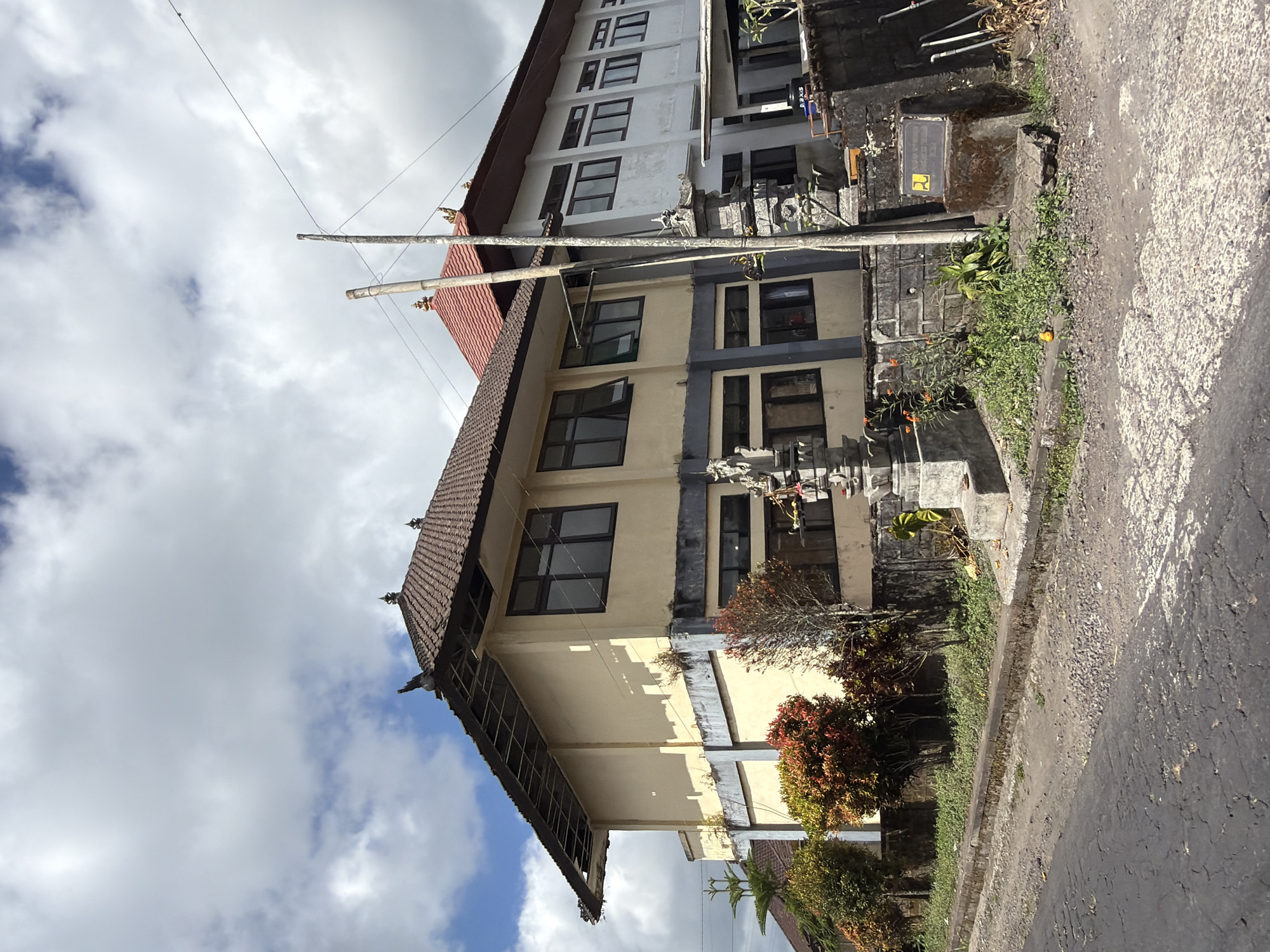 The image depicts two adjacent buildings in Kintamani, Indonesia, under a cloudy sky. The building on the left is a two-story structure with a light yellow facade, dark framed windows, and a traditional dark tiled roof with ornate finials. The lower story is accented with a blue band. To the right of this building, a white, multi-story building with a red tiled roof is partially visible, featuring numerous dark framed windows.
In the foreground, a street with a cracked asphalt surface runs along the side of the buildings. A low concrete curb separates the street from a strip of overgrown greenery and some decorative stone pillars. One pillar, intricately carved, stands prominently in front of the yellow building. In front of the white building, a dark stone structure holds a sign with a yellow logo.
The area appears to be an urban or semi-urban setting, possibly a public institution or commercial property. The vegetation consists of small trees with reddish-orange foliage and scattered green plants. Two utility poles are visible, one made of bamboo and another of metal, with electrical wires extending across the frame. The overall scene is an outdoor daytime view.