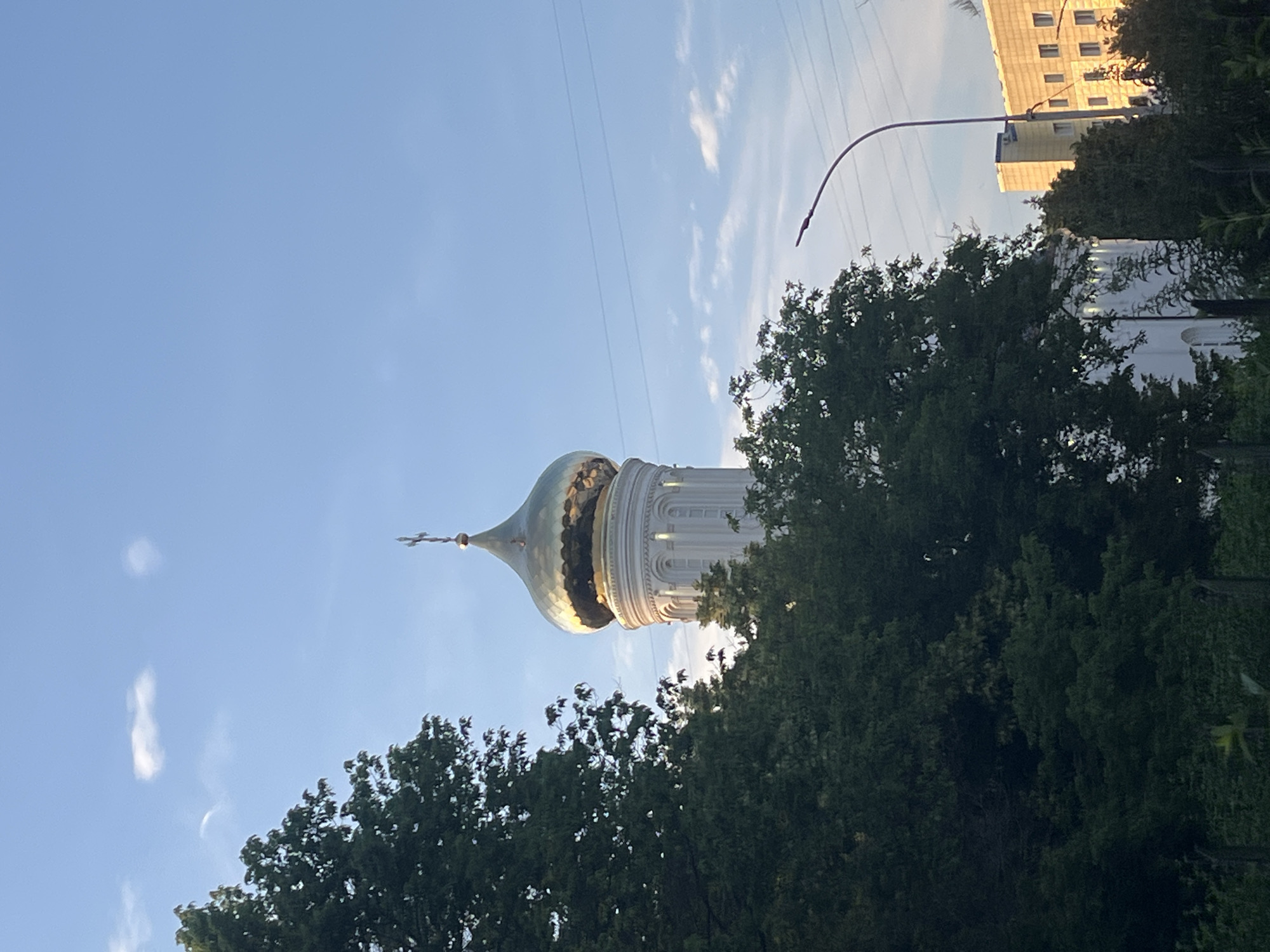 The image shows the dome of an Orthodox church, likely located in Krasnodar, Russia. The dome is golden and topped with a cross. The church tower is white and ornate, with arched windows. Surrounding the church are dense green trees and some buildings in the background. A street lamp is visible on the right side of the image. The sky is blue with scattered white clouds, suggesting it is daytime. There are no people visible in the image.