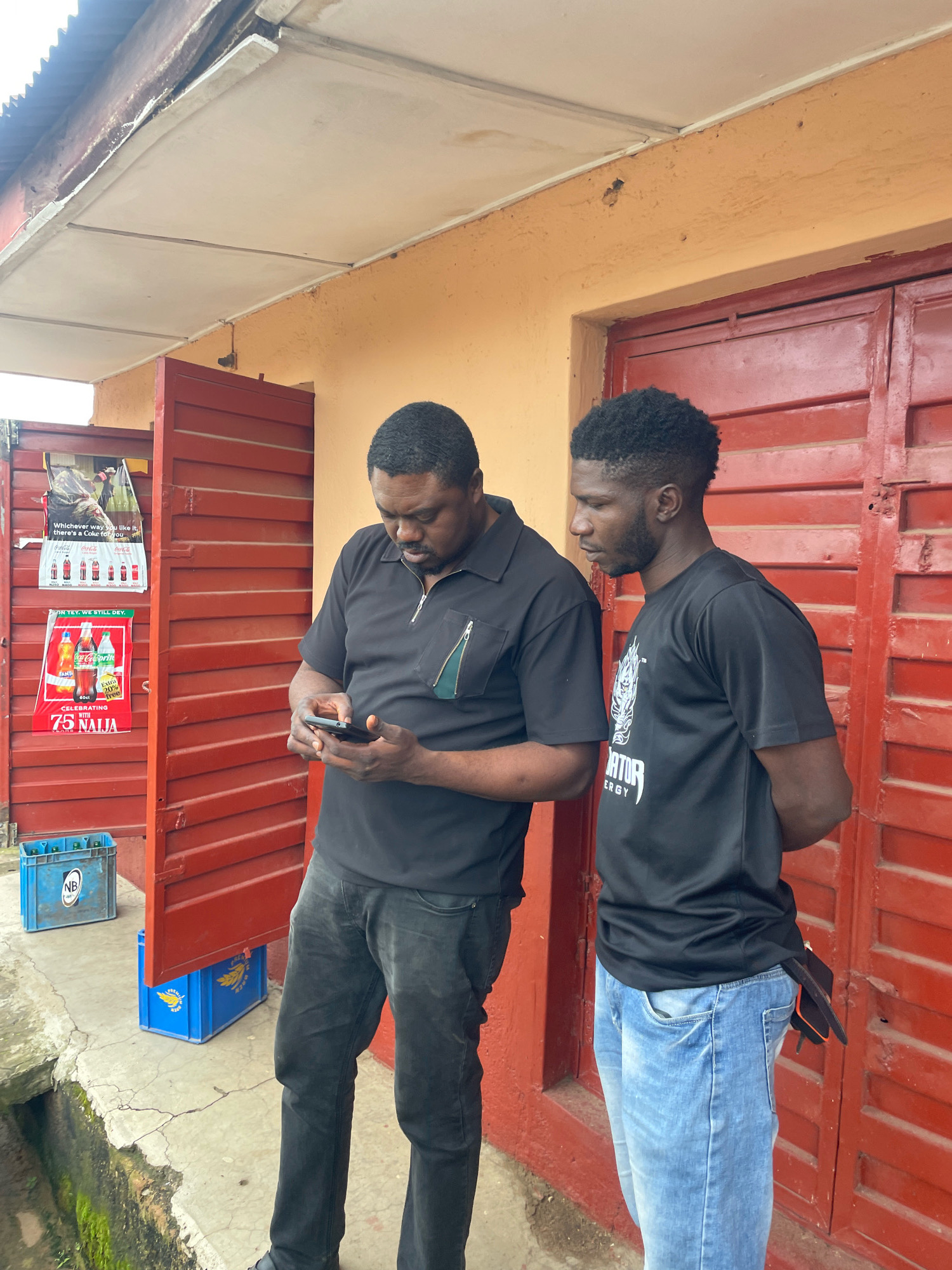 The image shows two men standing outside a building with red metal doors in what appears to be a market or commercial area in Jos, Nigeria. The man on the left, who is heavier set, is wearing a black polo shirt and dark grey jeans. He is looking down at his mobile phone, which he holds in his hands. The man on the right, who is slimmer with dark skin and short, curly hair, is wearing a black t-shirt with a graphic print and light blue jeans. He is standing with his hands behind his back, looking in the same direction as the other man, seemingly engaged in a conversation or observing something together. Behind the man on the left, there is a red metal door and a poster advertising Coca-Cola products. In front of the doors, there are blue plastic crates. The building has an orange-colored wall and a corrugated metal roof extending over the entrance. The ground is concrete with some visible cracks and moss growing in the lower left corner, suggesting an outdoor, possibly somewhat weathered, urban environment.
