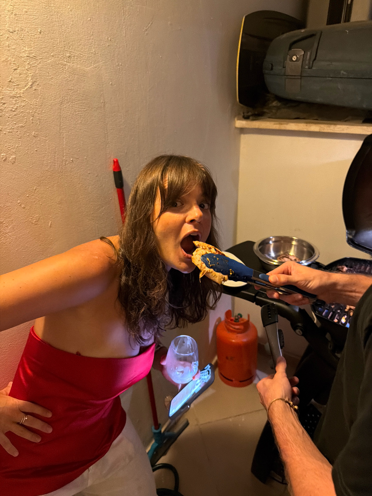 A woman with dark brown hair and bangs, wearing a strapless red top and white pants, is in the process of eating from a serving tong. Her mouth is open, and she is looking at the camera. A person's arm, wearing a bracelet, is holding the tongs and offering the food to the woman. In the background, there is a grill, a metal bowl, an orange gas tank, and a wall with a red cleaning tool leaning against it. The scene appears to be indoors or on a patio, possibly in Tel Aviv, Israel.