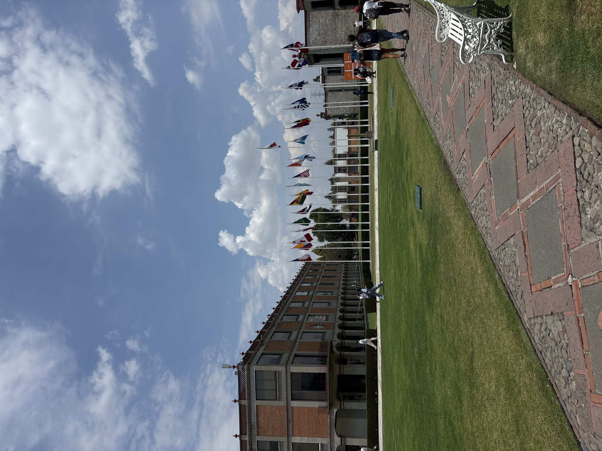 The image shows a view of the Universidad de las Américas Puebla (UDLAP) campus in San Andrés Cholula, Mexico. The camera is positioned along a brick and stone pathway bordered by a green lawn. A line of flagpoles, each flying a different national flag, stands parallel to the path. A long, multi-story brick building with windows and arches runs along the other side of the lawn. Several people are walking along the path, and a white bench is visible at the edge of the path. The sky is blue with scattered clouds.