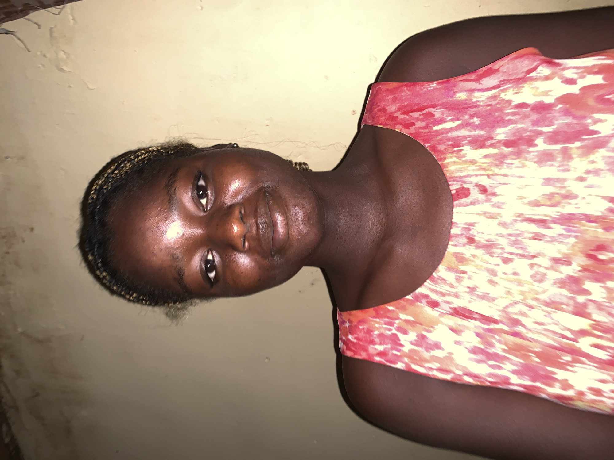A young woman with dark skin and braided hair is wearing a light-colored dress with reddish-pink patterns, posed indoors. The background shows a wall with a light-yellow tint, likely within a building in Jos, Nigeria.