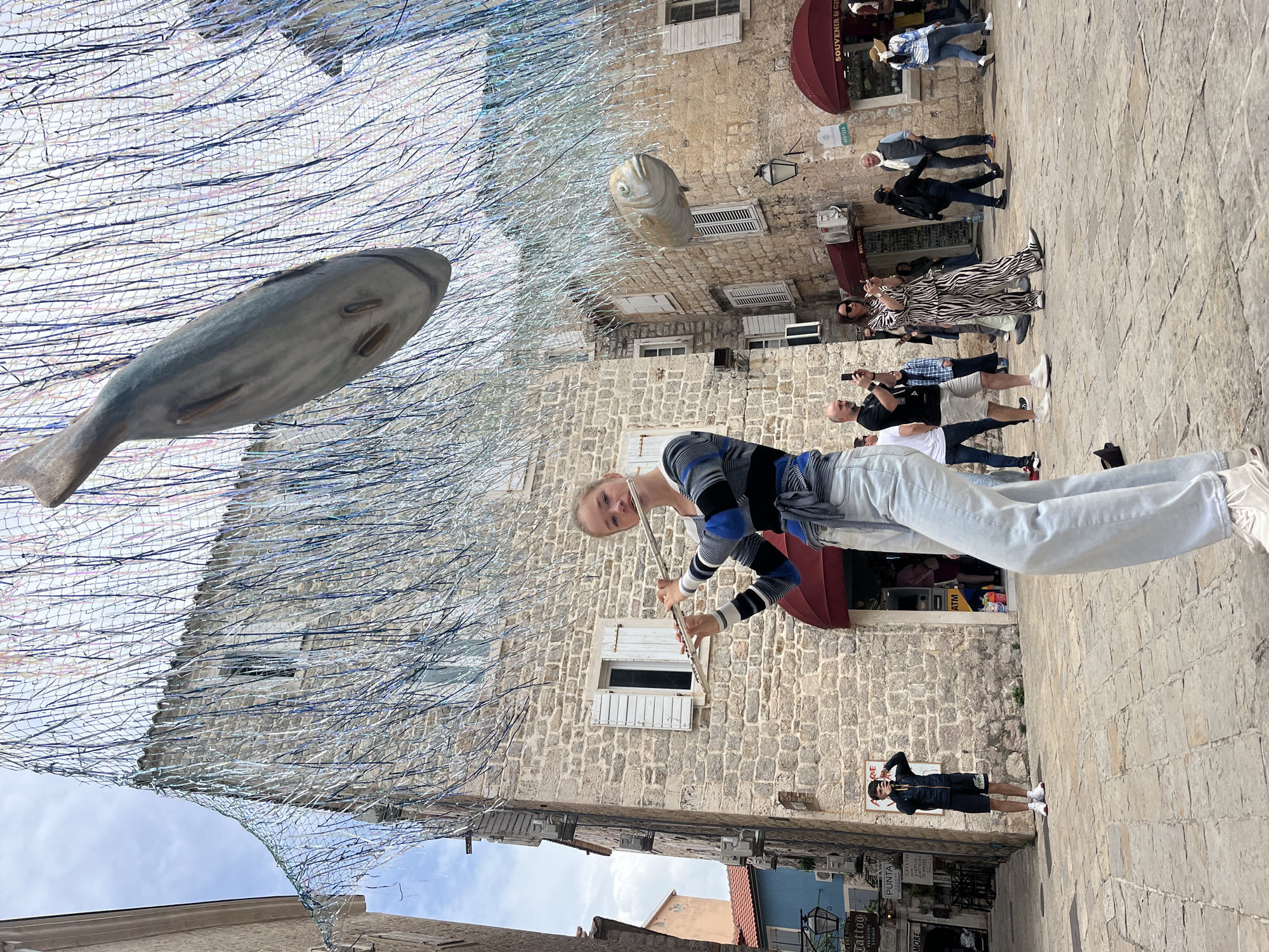 The image depicts a street scene in Budva, Montenegro, where the ancient stone architecture is prominent. Overhead, a decorative installation of large fish sculptures hangs suspended within a shimmering, net-like structure. In the foreground, a young woman stands playing a flute, positioned in front of one of the stone buildings. Several people are visible in the background, walking and observing the scene. The street is paved with stone blocks. Signage on buildings indicates businesses, including a souvenir shop and a tattoo studio. The sky is partly visible between the buildings and the overhead art installation.