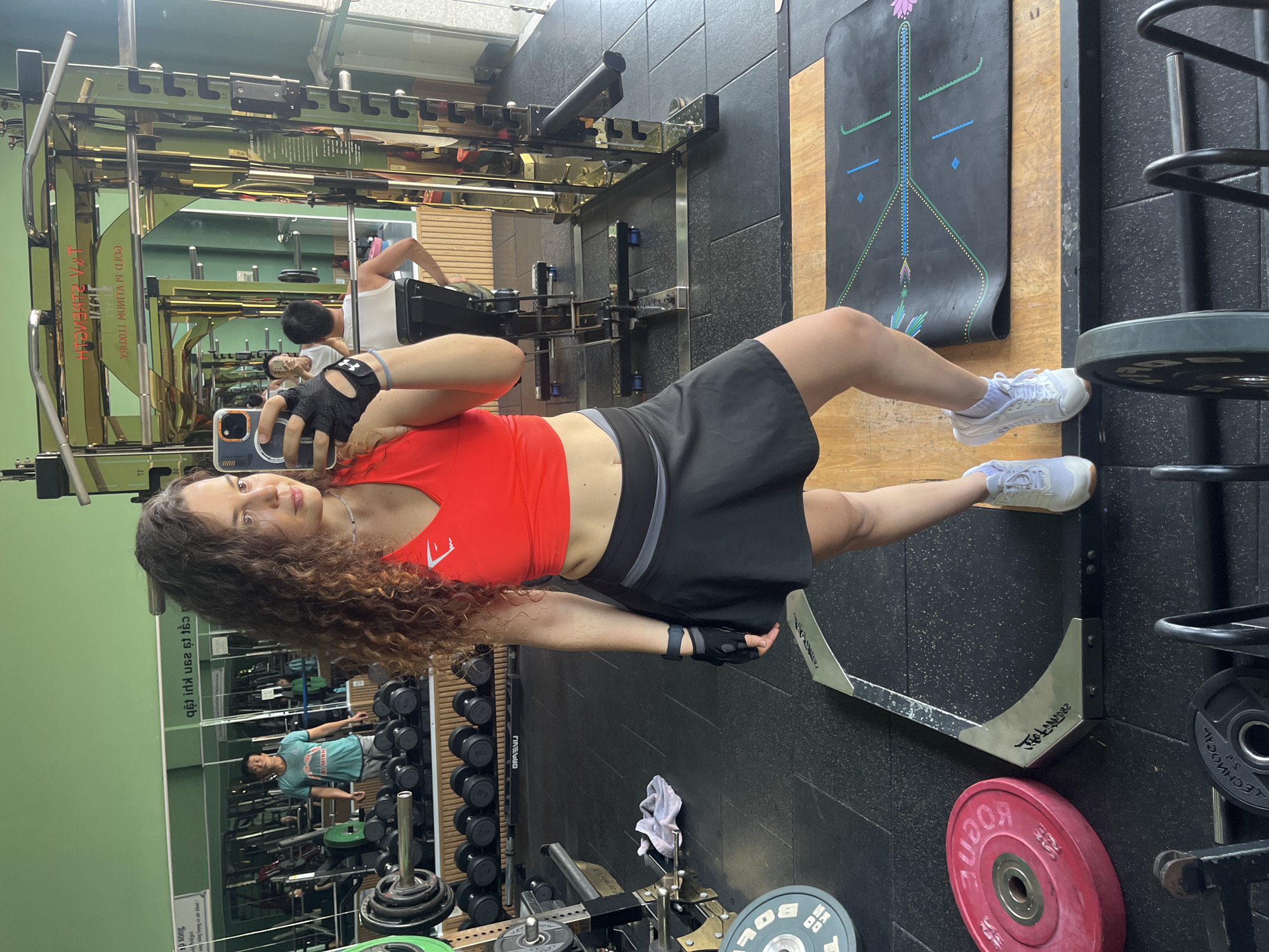 A woman with curly brown hair is taking a mirror selfie in a gym located in Phan Thiết, Vietnam. She is wearing a red sports bra, a black skirt, black workout gloves, and white sneakers. The gym appears to be well-equipped with weight racks, dumbbells, and weight plates. In the background, a man in a blue t-shirt is visible, also in the gym. The walls of the gym are painted green, and mirrors line one wall, reflecting the gym equipment and the people within. The floor is made of black rubber mats. A yoga mat with a floral design is visible on the floor in the foreground.