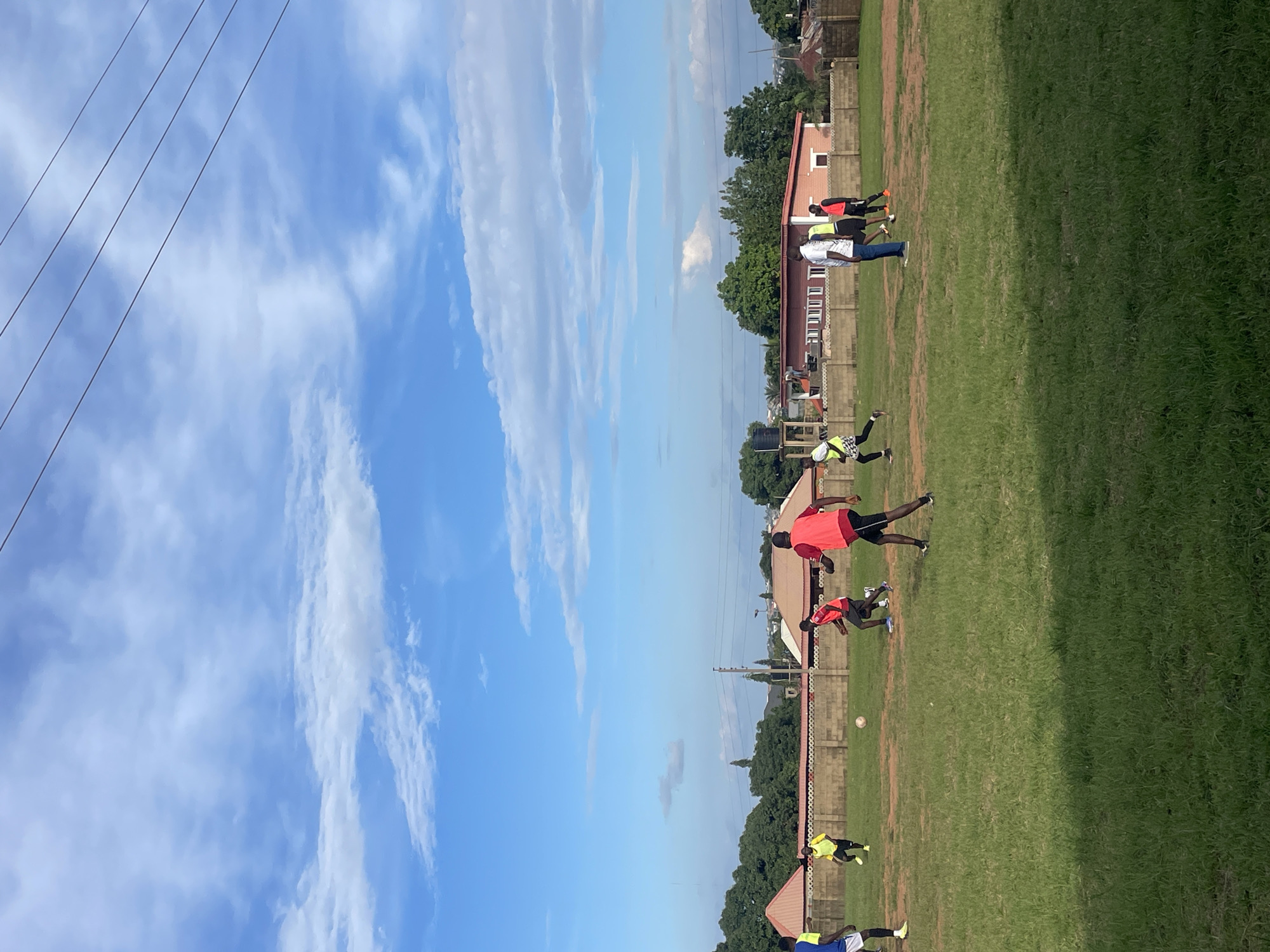 This image depicts a group of young men engaged in a soccer match on a grassy field, likely in Jos, Nigeria, given the context. The sky above is a vibrant blue, adorned with scattered white clouds, suggesting fair weather. In the background, residential buildings with red-tiled roofs and a low, beige-colored wall can be observed, interspersed with lush green trees. Several players are actively participating in the game, some in red jerseys, others in yellow vests, and one in a blue and white outfit. A soccer ball is visible on the field, indicating the game is in progress. The overall scene is one of youthful energy and outdoor activity within a semi-urban environment.