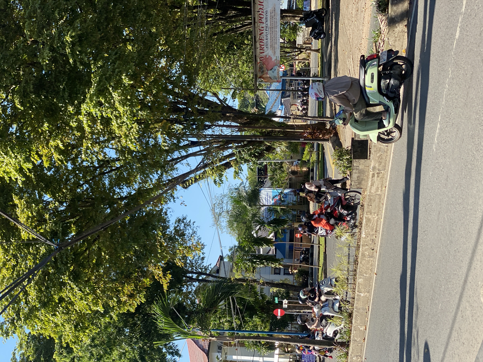 The image depicts a street scene in Banjarbaru, Indonesia, under a clear blue sky. Lush green trees line the side of the road, casting dappled shadows on the asphalt. In the foreground, a person rides a light green scooter, wearing a brown headscarf and dark pants. The scooter has a license plate visible.
Further down the street, several other individuals on motorcycles and scooters are present. Some are parked, while others are in motion. One white scooter is occupied by two people. A person in a bright orange shirt is riding a red scooter. A woman in a beige headscarf and dark clothing walks on the sidewalk.
Buildings with red tiled roofs are visible in the background, along with palm trees and smaller ornamental plants. A banner with Indonesian text, including "WAROENG PODJO," is displayed on the right side of the scene, suggesting a commercial establishment. Traffic signs, including a red circular sign with a white horizontal bar, are also present. The overall impression is a bustling urban environment on a sunny day.