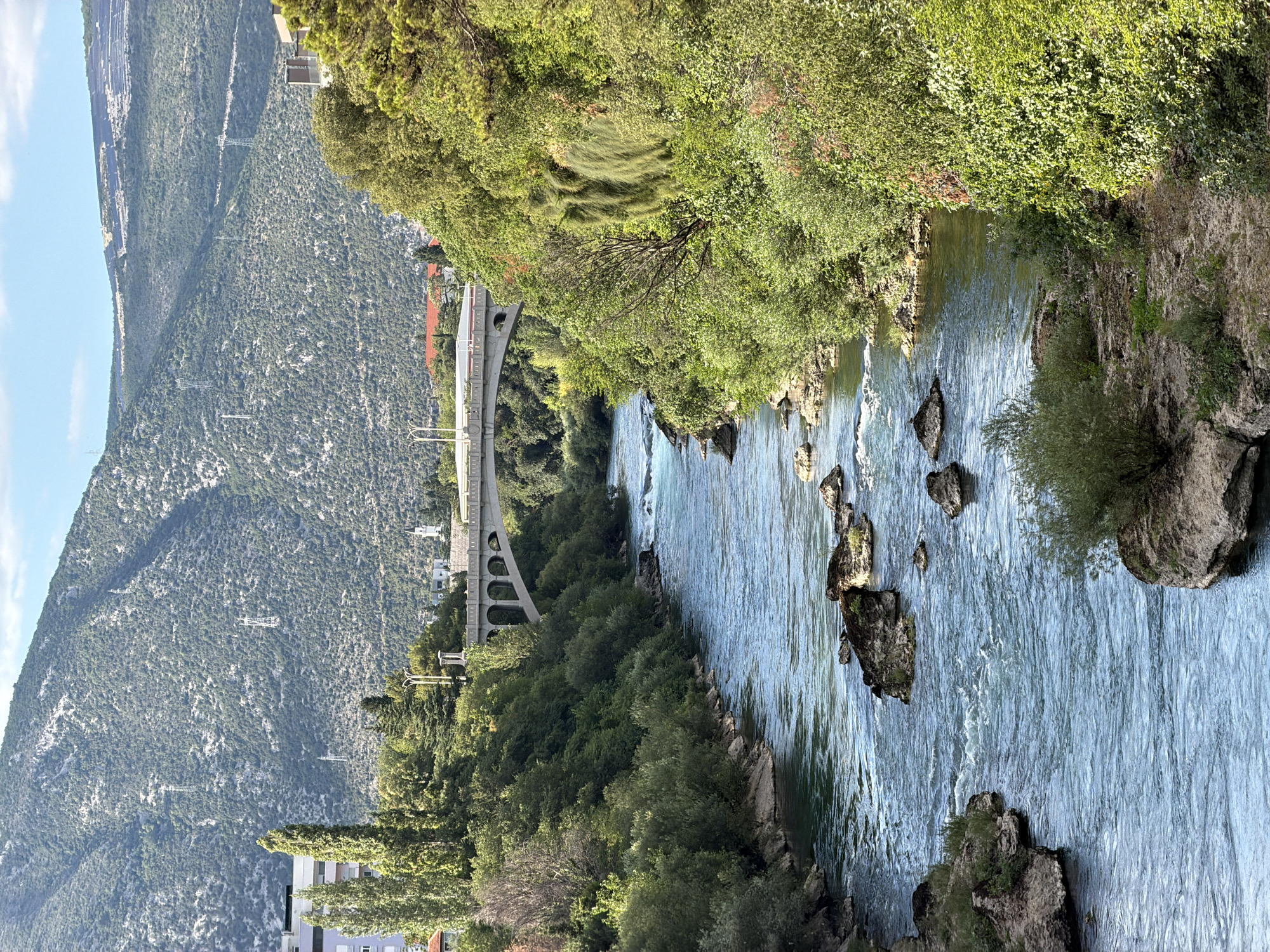 A wide shot captures a natural river flowing through an urban landscape in Mostar, Bosnia and Herzegovina. The river, a vibrant turquoise, is partially obstructed by scattered grey rocks, with visible white-capped rapids in the foreground. The banks are densely lined with lush green trees and shrubs, with some rocky outcrops. In the mid-ground, a modern concrete bridge with multiple arches spans the river. Beyond the bridge, glimpses of buildings and more greenery are visible, including tall, slender trees. In the background, a steep, verdant mountain rises, its slopes covered in dense foliage and dotted with visible power line pylons and transmission towers. The sky is a clear, bright blue with a few scattered white clouds. The scene is illuminated by bright sunlight, suggesting it is daytime. There are no people or animals visible in the frame.