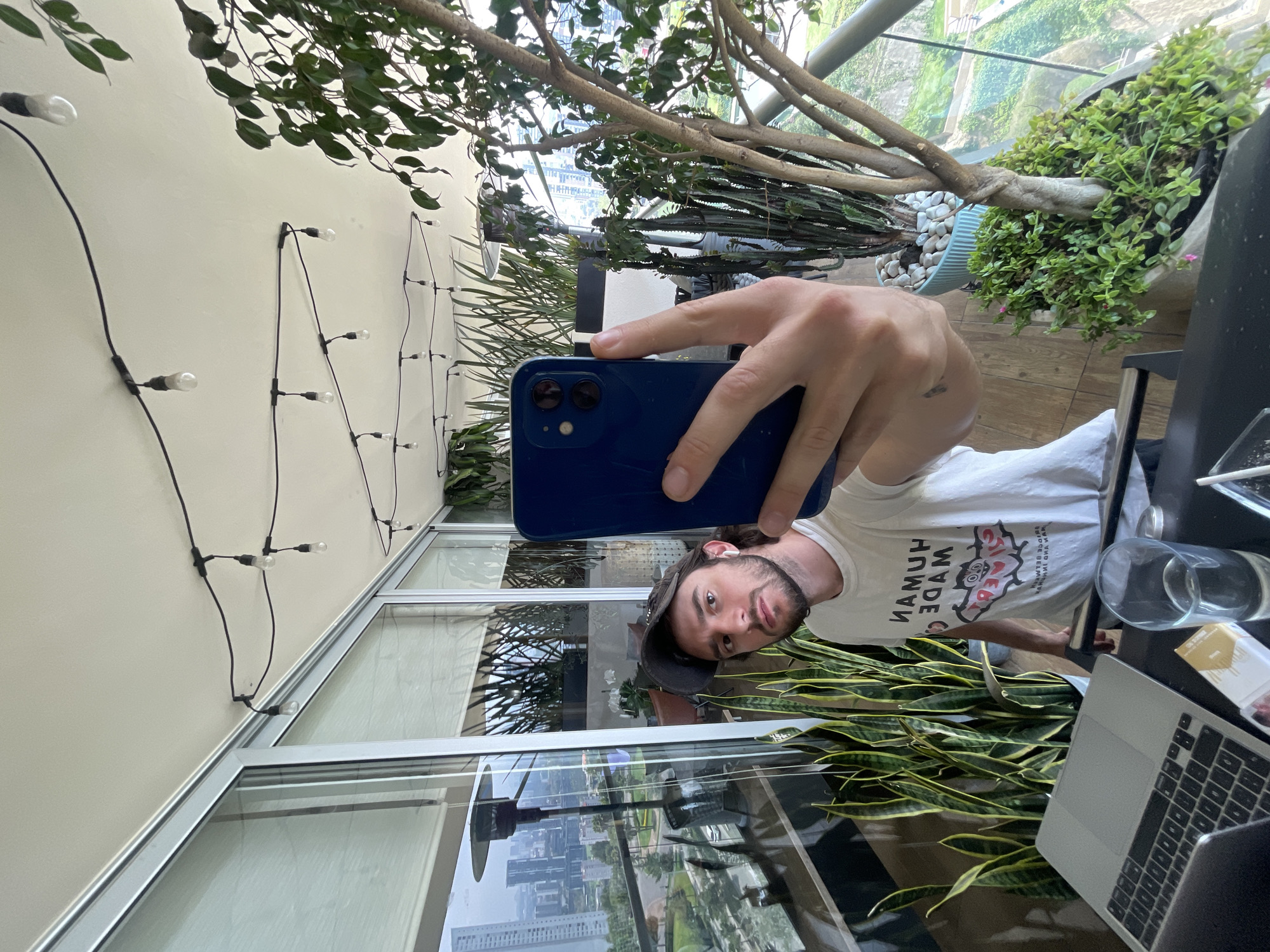 A person is taking a selfie with a blue smartphone in what appears to be a modern, plant-filled indoor space. The person has a beard, and is wearing a t-shirt and a baseball cap.
The background includes:
-Large windows reflecting a cityscape. The city is Huixquilucan de Degollado, Mexico.
-Various potted plants, including snake plants and other greenery.
-String lights along a wall.
-A laptop, a glass of water, and a pack of cigarettes on a dark table.