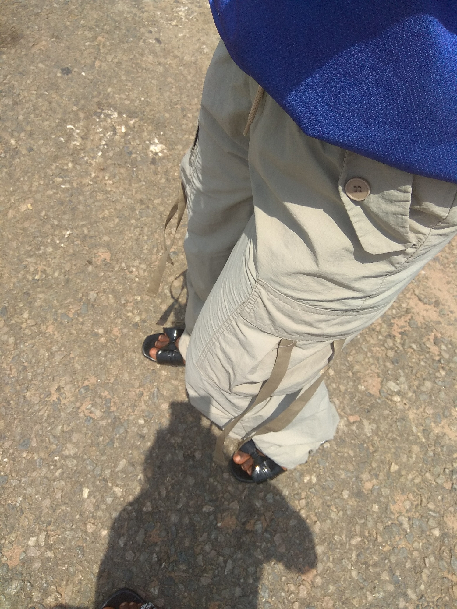 The image shows a close-up view of a person's lower body, standing on a paved surface that appears to be a road or walkway. The person is wearing light-colored cargo pants and black sandals. The pants have multiple pockets and straps, and a prominent button is visible on the right pocket. The person is also wearing a blue shirt, the top portion of which is visible. A shadow of the person is cast on the ground. The ground is covered with small stones and gravel, typical of a road surface in Jalingo, Nigeria. The lighting suggests it is daytime, with the sun casting a strong shadow.