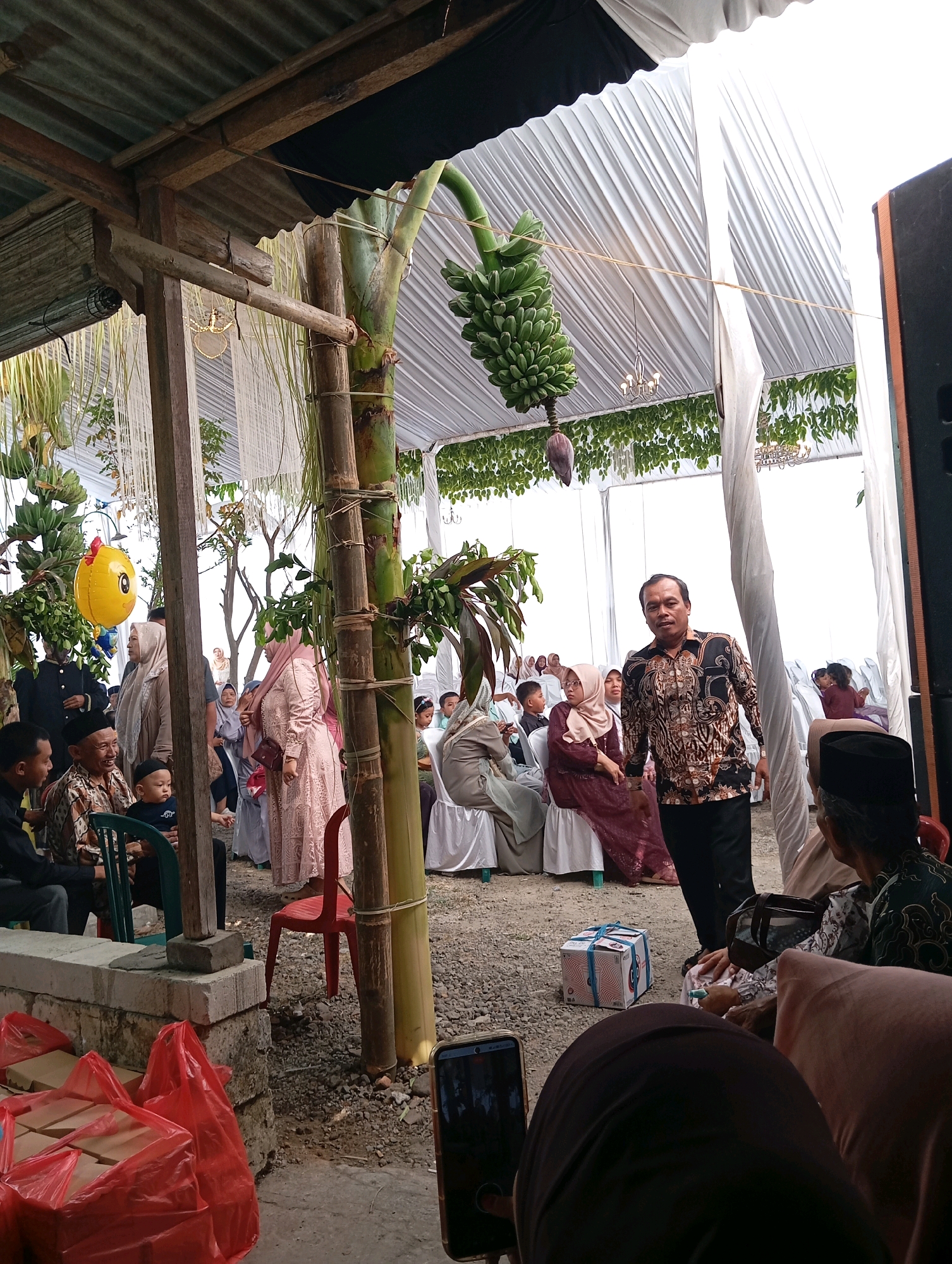 The image depicts an outdoor event, likely a wedding or celebration, taking place under a white tent in Ngampel, Indonesia. The ground is covered in gravel. A large banana bunch hangs from a stalk near the center, and a yellow balloon in the shape of a cartoon character is visible on the left. A man in a patterned shirt and dark pants is standing and looking towards the camera, possibly addressing the guests. Several people are seated on chairs, some wearing traditional attire including headscarves. Red plastic bags filled with items are stacked on the lower left, and a box tied with blue ribbon sits on the ground. A cell phone is visible in the foreground, suggesting someone is recording or taking pictures of the event. The overall atmosphere appears to be festive and social.