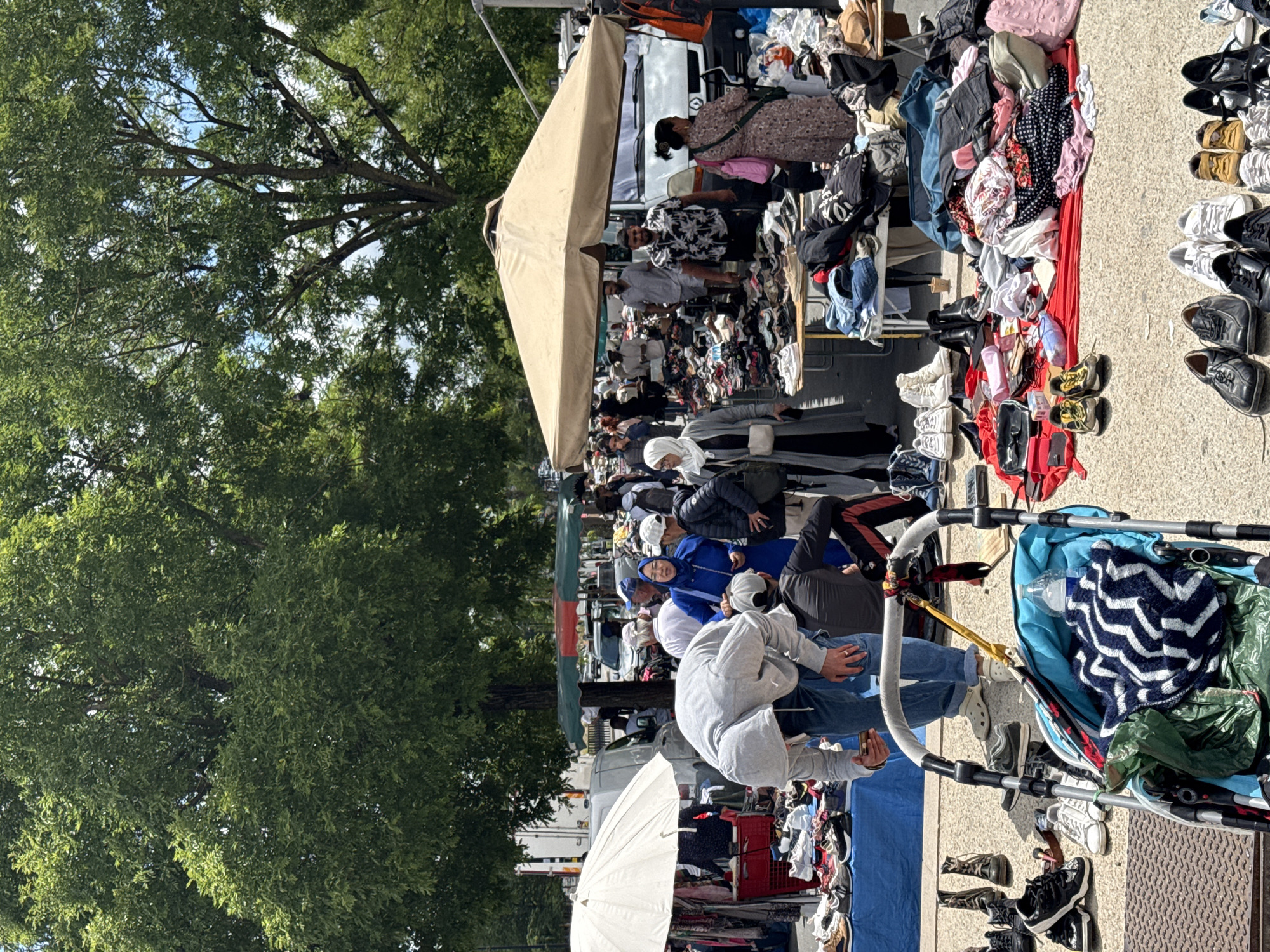 This is a street market scene, likely in Bordeaux, France, given the style of the market and the general ambiance. The foreground is dominated by a close-up of a baby stroller with a blue and white zigzag blanket and a green bag inside. To the left of the stroller, a collection of shoes is displayed on the ground, including black boots, sneakers, and loafers. Behind the stroller, a man in a grey hoodie and jeans is bent over, looking at his phone. A woman in a bright blue outfit and headscarf stands nearby, facing him.
Further back, the market unfolds with stalls displaying clothing and shoes. Several people are browsing the merchandise. A large beige umbrella shades a section of the market where shoes are laid out on tables. A white van is parked behind some of the stalls. In the background, mature trees with dense green foliage form a canopy over the market area. The ground is paved with asphalt or concrete. The overall scene depicts a lively outdoor market on a bright, sunny day.