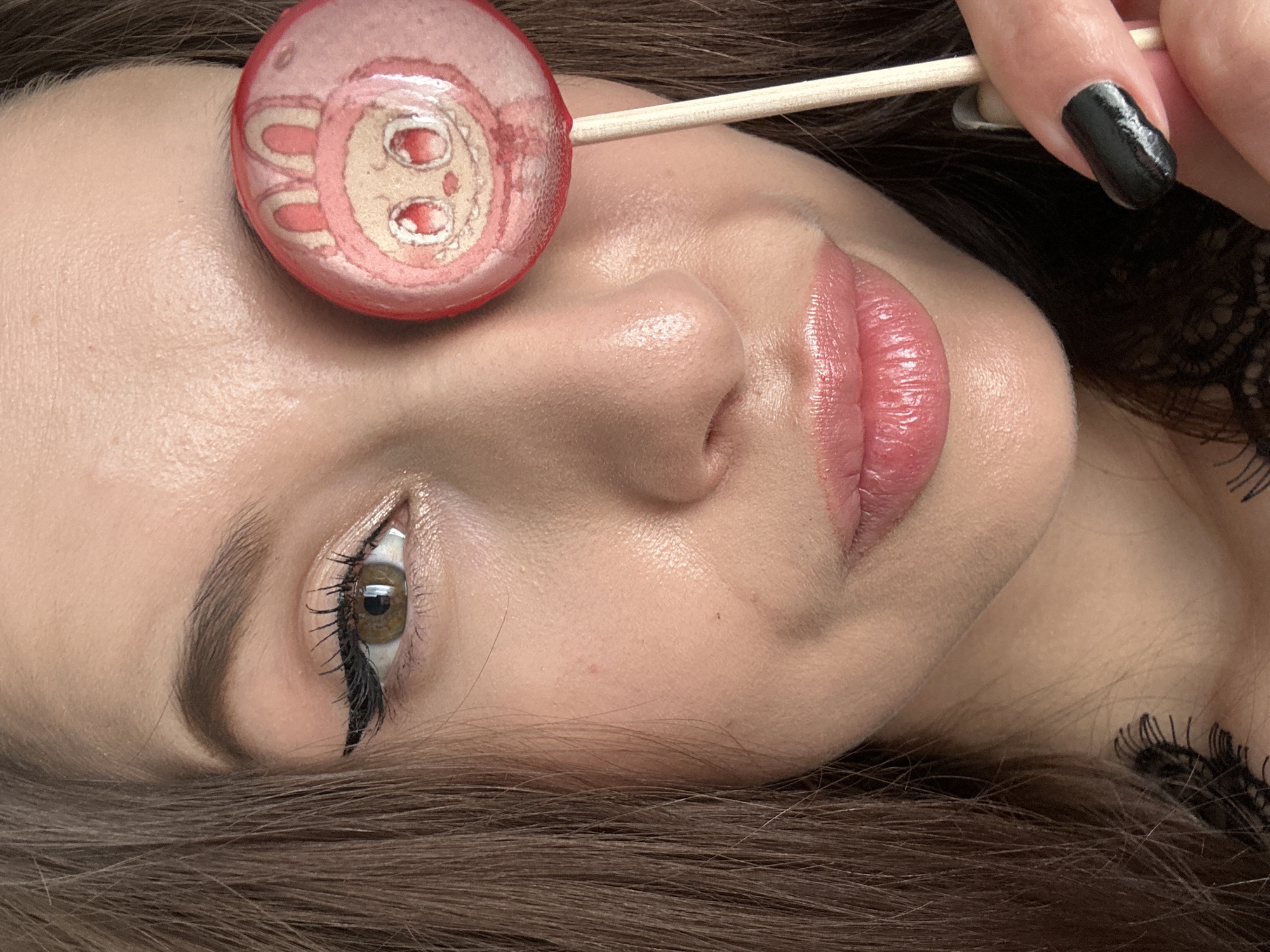 This close-up portrait features a woman with brown hair, lying down, likely indoors in Yekaterinburg, Russia. Her face, primarily the right side, fills the frame. She has a neutral and calm expression, with her right eye open and looking upwards, revealing a light brown iris. Her eyebrows are well-defined, and she wears black eyeliner and mascara. Her lips are full and adorned with a pink, glossy lipstick. Her skin appears smooth, with a subtle sheen, particularly on her forehead, nose, and cheekbone.
A round, flat lollipop with a light pink base and a cartoonish design of a character's face (resembling a rabbit or similar animal with large eyes and ears) is placed on her forehead, near her temple. A thin, light-colored wooden stick extends from the lollipop, across her face. A hand with a black-painted fingernail is seen holding this lollipop stick near the woman's temple. Part of a black, delicate lace garment, possibly a bra strap or top, is visible on the right side of the image, resting on her shoulder or neck area. The overall activity appears to be a posed photograph, possibly for a beauty or lifestyle context, with the lollipop adding a whimsical or playful element. There is no visible text in the image.