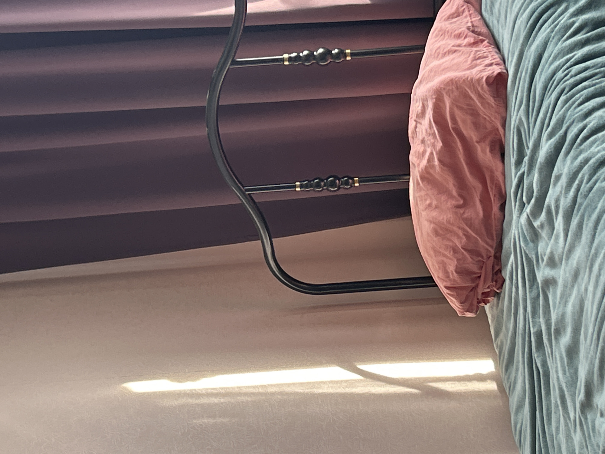 The image depicts a close-up view of a bed with blue-green bedding and a crumpled pink pillow. The bed is made of black metal with decorative gold and black accents on the headboard. In the background, there are two curtains, one a light beige and the other a deep purple. A shaft of bright sunlight falls on the beige curtain, casting a shadow on the bed. The scene appears to be indoors, possibly in a bedroom. There is no visible indication of the location being in Moscow, Russia.