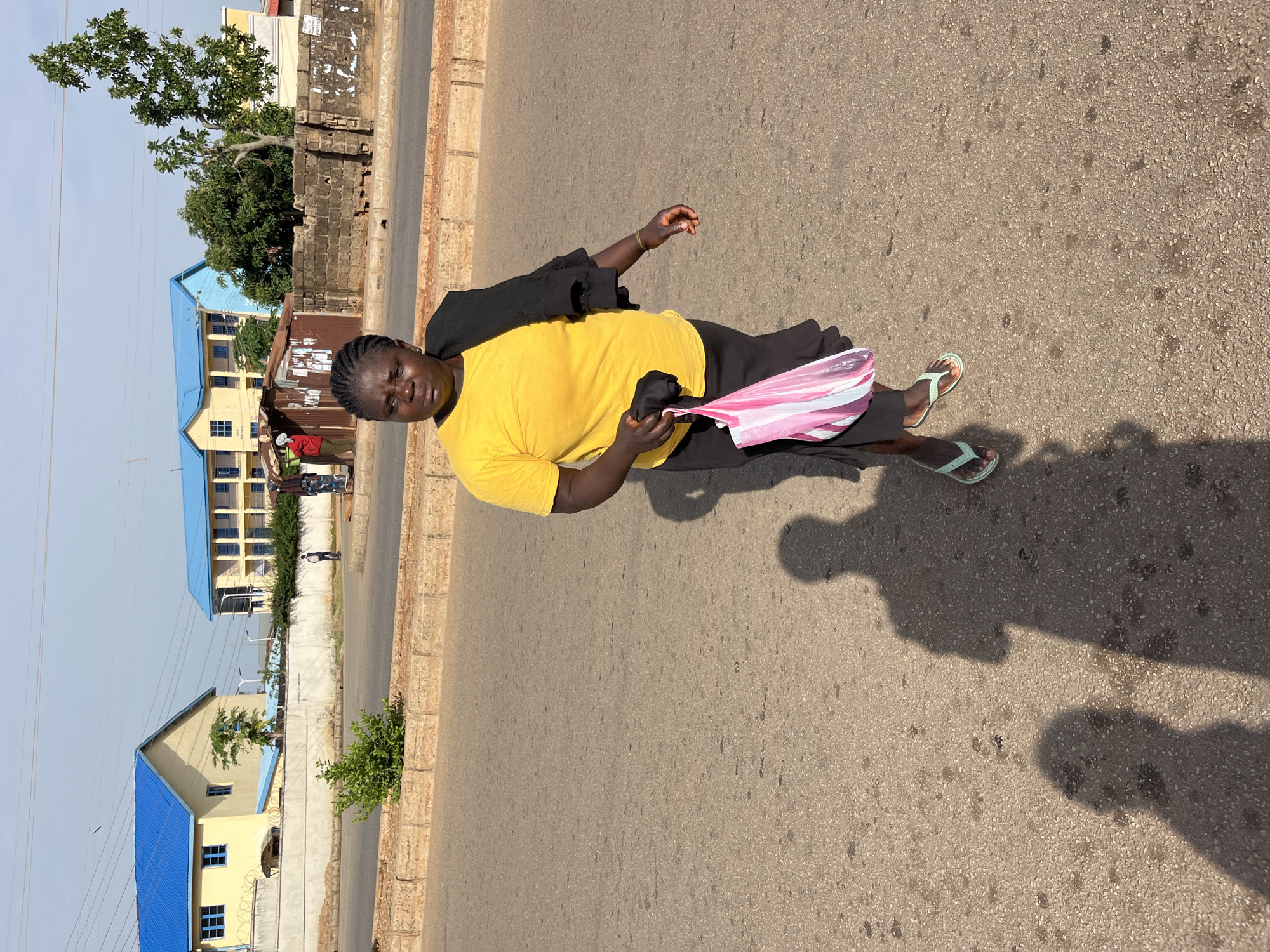 A woman stands on a paved road, holding a pink and white striped object that resembles a folded umbrella or bag. She is wearing a bright yellow t-shirt and a black covering over her shoulders. Her hair is braided. She is also wearing light green flip-flops. Her shadow is cast on the road.
In the background, there are buildings with blue roofs, some appearing to be schools or administrative buildings. A tree with green foliage is visible, and a brick wall runs along the side of the road. The scene appears to be outdoors during the day, with clear skies. The overall impression is a casual street scene in a populated area. The location context is Bukuru, Nigeria.