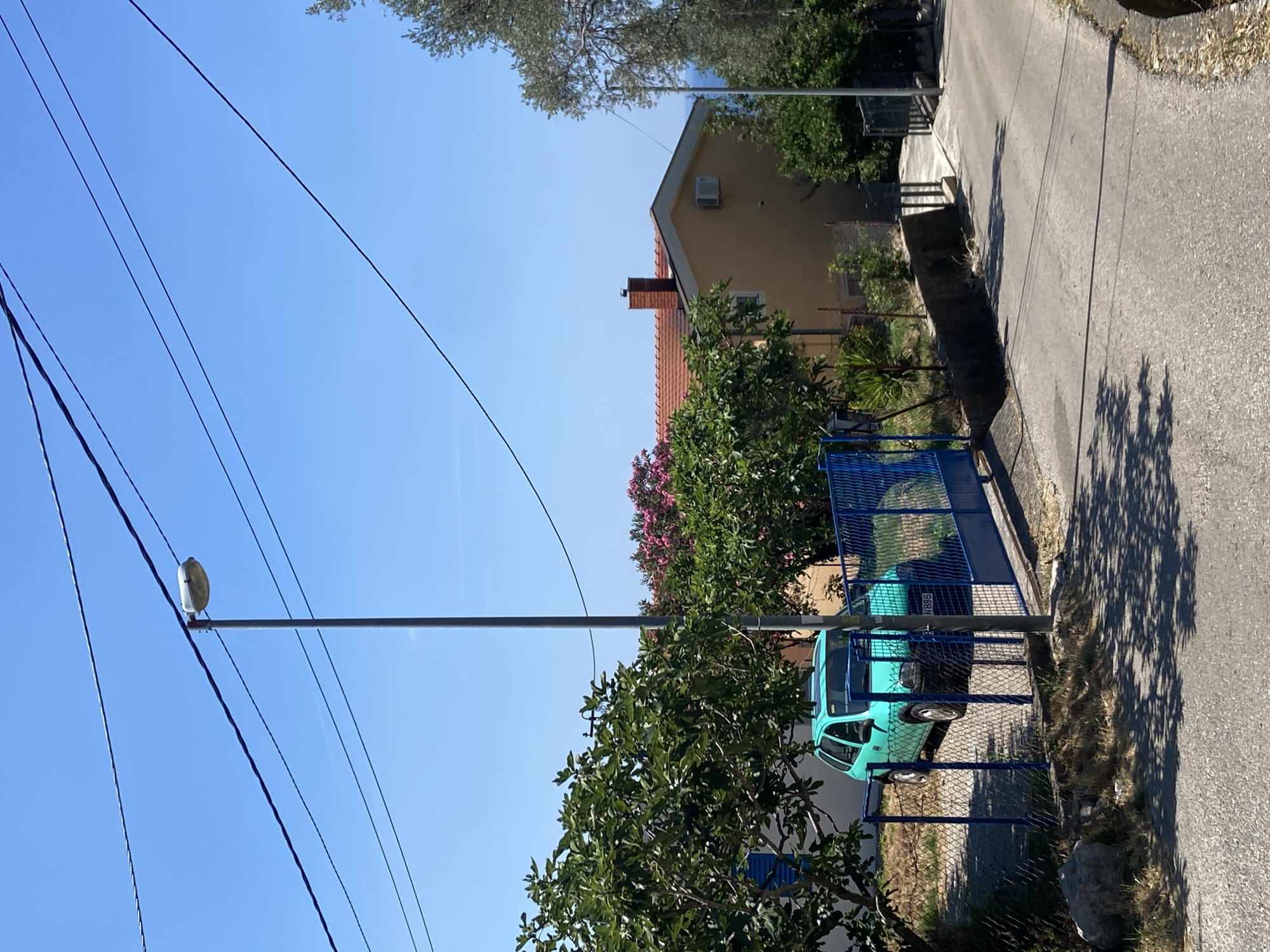 This image depicts a sunny day in Šušanj, Montenegro, with a clear blue sky overhead. In the foreground, a grey asphalt road curves to the right, shaded by dappled sunlight filtering through unseen trees. To the left of the road, a bright turquoise car is parked behind a blue metal fence. The car appears to be a small hatchback, possibly a late 1990s or early 2000s model, with a license plate displaying "N886". Adjacent to the car and fence is a large tree with lush green foliage.
Beyond the fence and car, a light yellow house with a terracotta tiled roof is visible. The house has a red brick chimney and a white air conditioning unit mounted on the exterior wall. Pink flowering bushes are planted in front of the house, adding a splash of color. Further to the right, an olive tree with silvery-green leaves stands tall, its branches reaching towards the sky. The background reveals a glimpse of the Adriatic Sea in the distance. A tall metal pole with a street lamp stands prominently near the center of the image, with power lines crisscrossing the sky. The overall scene conveys a sense of a quiet residential area on a warm summer day.