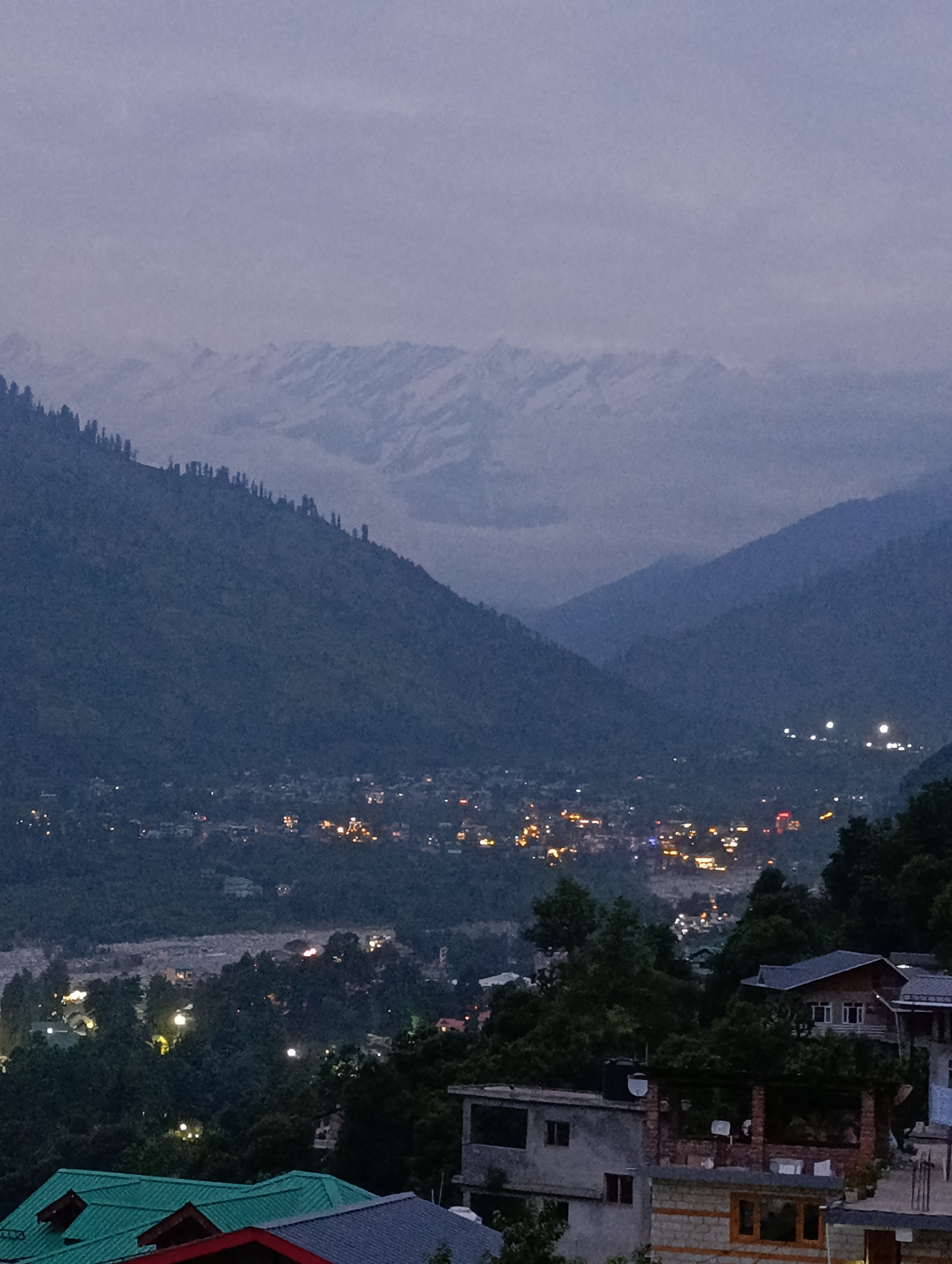 The image depicts a twilight view of a mountainous landscape with a town nestled in the valley. Snow-capped mountains are visible in the background, partially obscured by haze. The midground features a densely populated town illuminated by numerous lights, suggesting it is evening or night. Trees and darker forested hills surround the town and extend into the foreground. In the immediate foreground, several houses with pitched roofs, some green and some dark, are visible. One house in the mid-foreground has a dark tiled roof and multiple windows. Another house to its right is multi-storied with a stone facade. The overall atmosphere is serene and offers a glimpse of a settlement in a mountainous region.