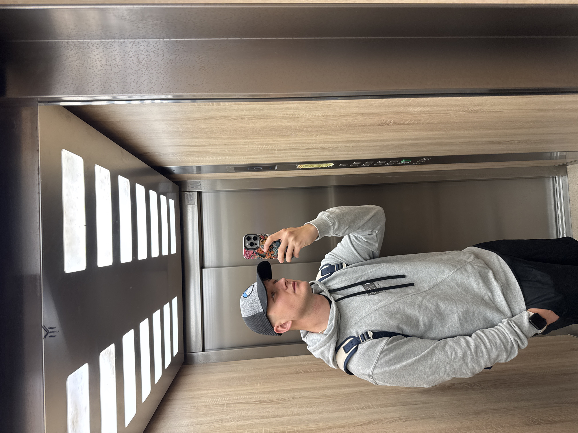 A young man, likely in Plovdiv, Bulgaria, is pictured inside an elevator, holding a smartphone to take a selfie. He is wearing a light grey hooded sweatshirt, dark pants, a grey and black baseball cap featuring a logo, and a smartwatch on his left wrist. Visible backpack straps are over his shoulders. His smartphone, held up in his right hand, is adorned with a colorful, intricately patterned case.
The elevator interior showcases a contemporary design. The floor and ceiling are finished with light wood-patterned panels, while the walls are sleek metallic grey. On the left wall, a dark decorative panel features multiple vertical rectangular cutouts, illuminated from behind to create ambient lighting. A narrow control panel strip, featuring various buttons and indicator lights, including a green light and some yellow illumination, runs horizontally along the metallic wall just beneath the wooden ceiling. The man has a neutral expression, focusing on his phone.