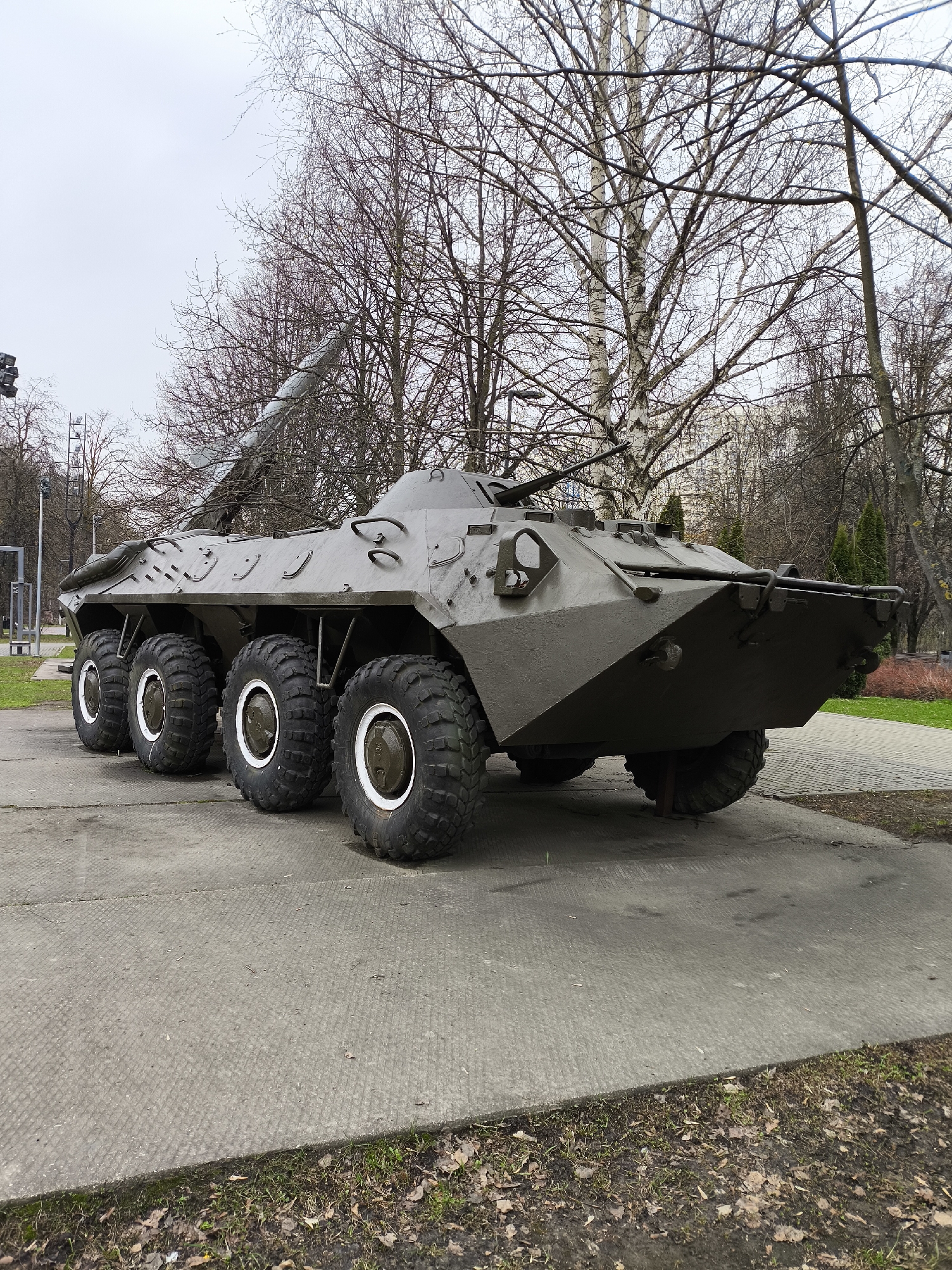 An olive-green, eight-wheeled armored vehicle, likely a BTR-series Armored Personnel Carrier, is displayed outdoors on a concrete slab. The vehicle has a rugged appearance with large, thick tires featuring white rims. Its hull is angled, and various hatches and fixtures are visible on its surface. A small turret with what appears to be a machine gun is mounted towards the front. The vehicle is stationary, serving as a monument or exhibit.
The setting is a park or public space, characterized by numerous deciduous trees, mostly bare of leaves, suggesting late autumn, winter, or early spring. A tall, slender white-barked tree, possibly a birch, stands prominently in the background. Through the branches, glimpses of distant buildings can be seen, indicating an urban or suburban environment. To the left, a tall metal structure with floodlights is visible, and behind the armored vehicle, a long, cylindrical object extends upwards at an angle, possibly a rocket or missile on display, or an art installation.
The ground around the concrete slab is covered with sparse grass, dirt, and fallen brown leaves. The sky is overcast, contributing to a muted, cool-toned ambiance. No people are present, and there is no discernible activity other than the static display of the military vehicle. No visible text is present in the image.