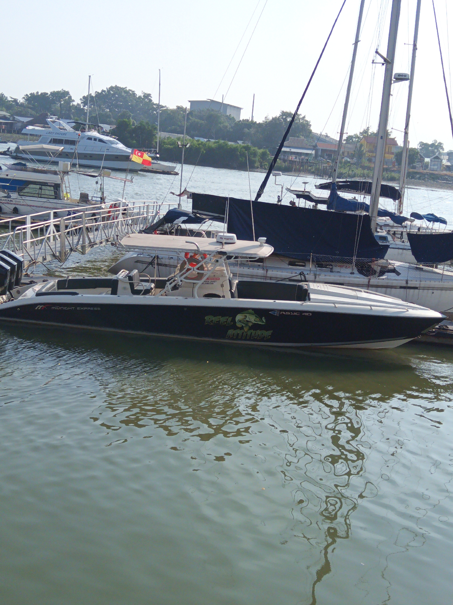 The image depicts a marina scene in Klang City, Malaysia, with several boats docked. In the foreground, a dark-hulled motorboat named "MIDNIGHT EXPRESS" with the name "FEEL ATTITUDE" emblazoned on its side is prominently featured. It has a white console and seating area. Behind it, a larger white yacht named "LADY THOESE" is visible, adorned with a Malaysian flag and another flag that appears to be the flag of Selangor. Further back, other boats, including sailboats, are moored. The water in the marina is calm with reflections of the boats and surrounding trees. The background shows a shoreline with vegetation and buildings, indicating an urban coastal environment. The sky is bright and overcast. There are no people visible in the image. The overall scene is a snapshot of a typical day at a Malaysian marina.