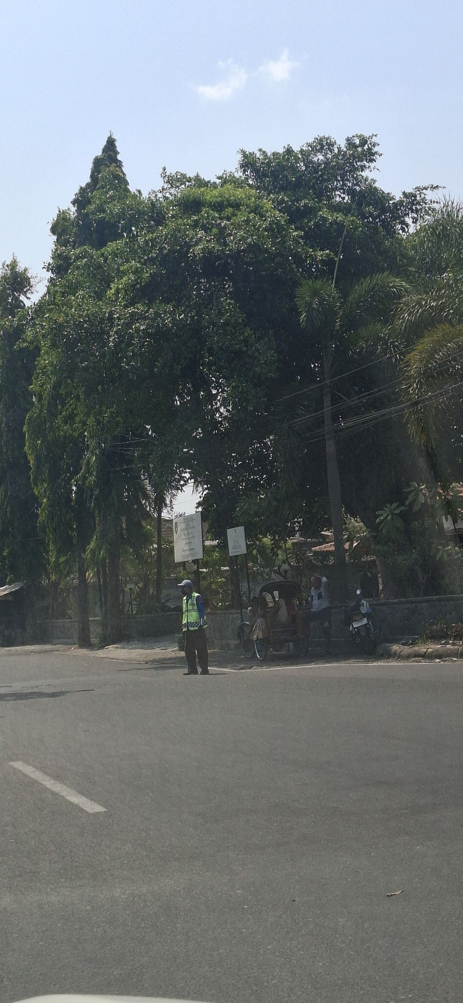 A man in a yellow safety vest and blue cap stands on the side of a road in Surakarta, Indonesia. He appears to be a traffic officer. Behind him, there is a three-wheeled vehicle and a motorcycle. The road is paved and has a white line marking. In the background, there are trees and buildings. The sky is clear and blue with a few clouds.