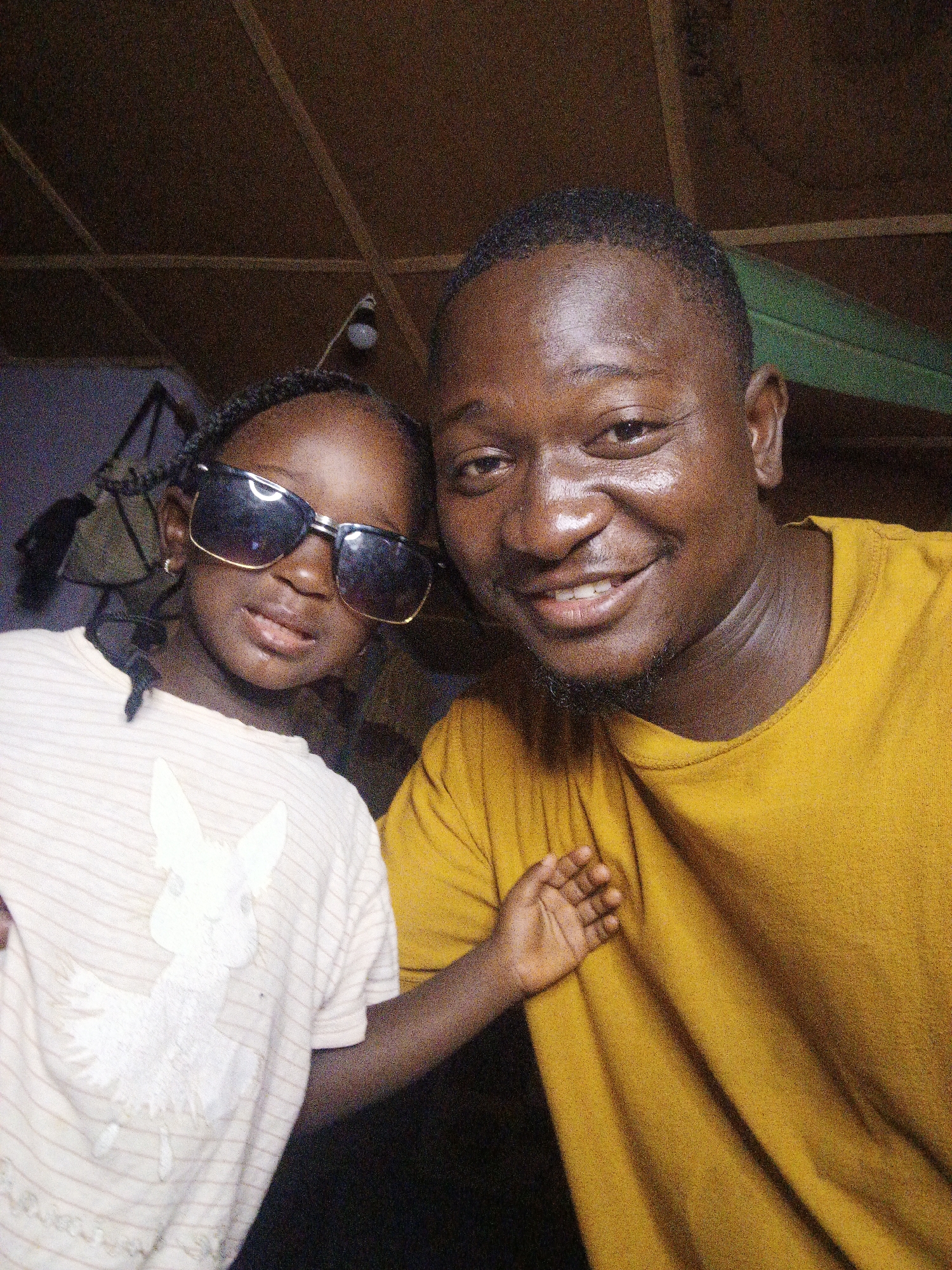 A man and a young girl are posing for a selfie. The man is on the right, wearing a yellow t-shirt and smiling. The girl is on the left, wearing a white t-shirt with a cartoon bunny design and large sunglasses. They are standing close together, with the man's arm around the girl. The background appears to be an indoor space with a wooden ceiling and some hanging objects. The lighting is dim, suggesting the photo was taken in low-light conditions.