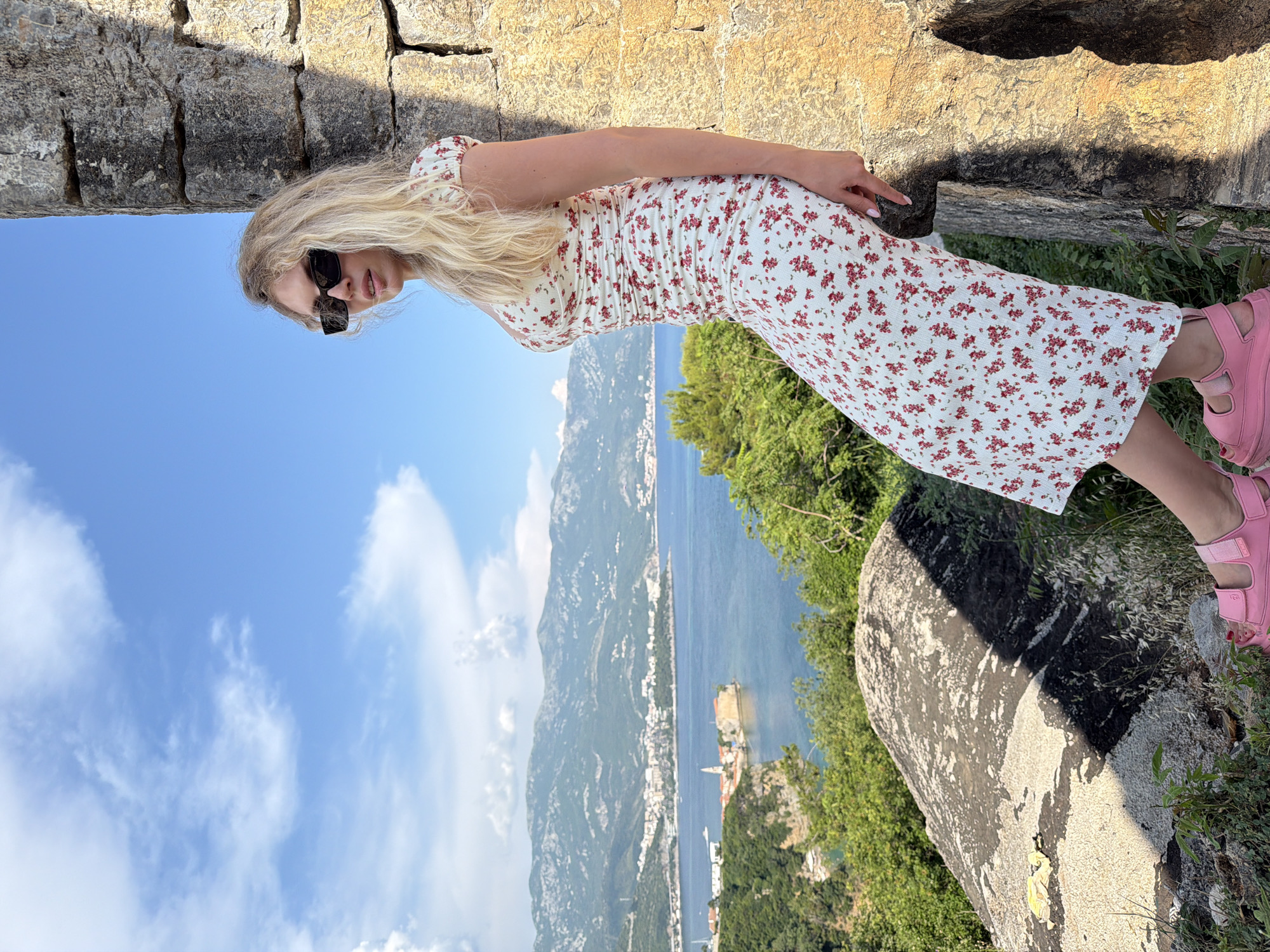 A blonde woman with sunglasses and a floral print dress leans against a stone wall. She is outdoors with a view of the Adriatic Sea and the city of Budva, Montenegro. The city is nestled between rolling green hills and the blue sea, with buildings and a church visible on a small peninsula. The sky is blue with scattered white clouds. The woman is wearing pink platform sandals.
