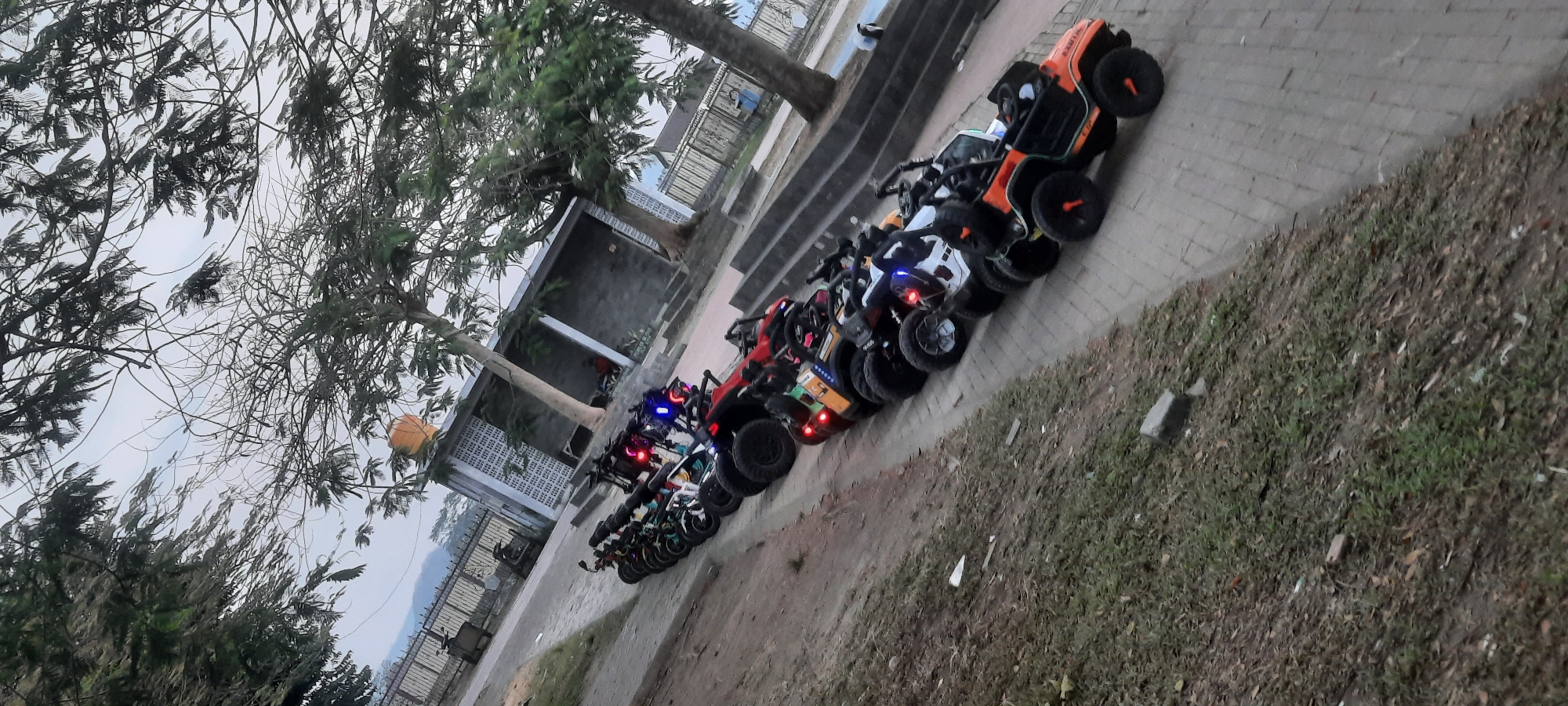 An outdoor, eye-level, vertical shot shows a line of parked quad bikes on a paved path next to a grassy slope. The quad bikes are various colors and appear to be children's ride-on toys. Some have blue LED lights illuminated. In the background, there is a low fence, a building with a tiled roof, and trees with sparse foliage. The sky is overcast. The location is the city of Kanigoro, Indonesia.