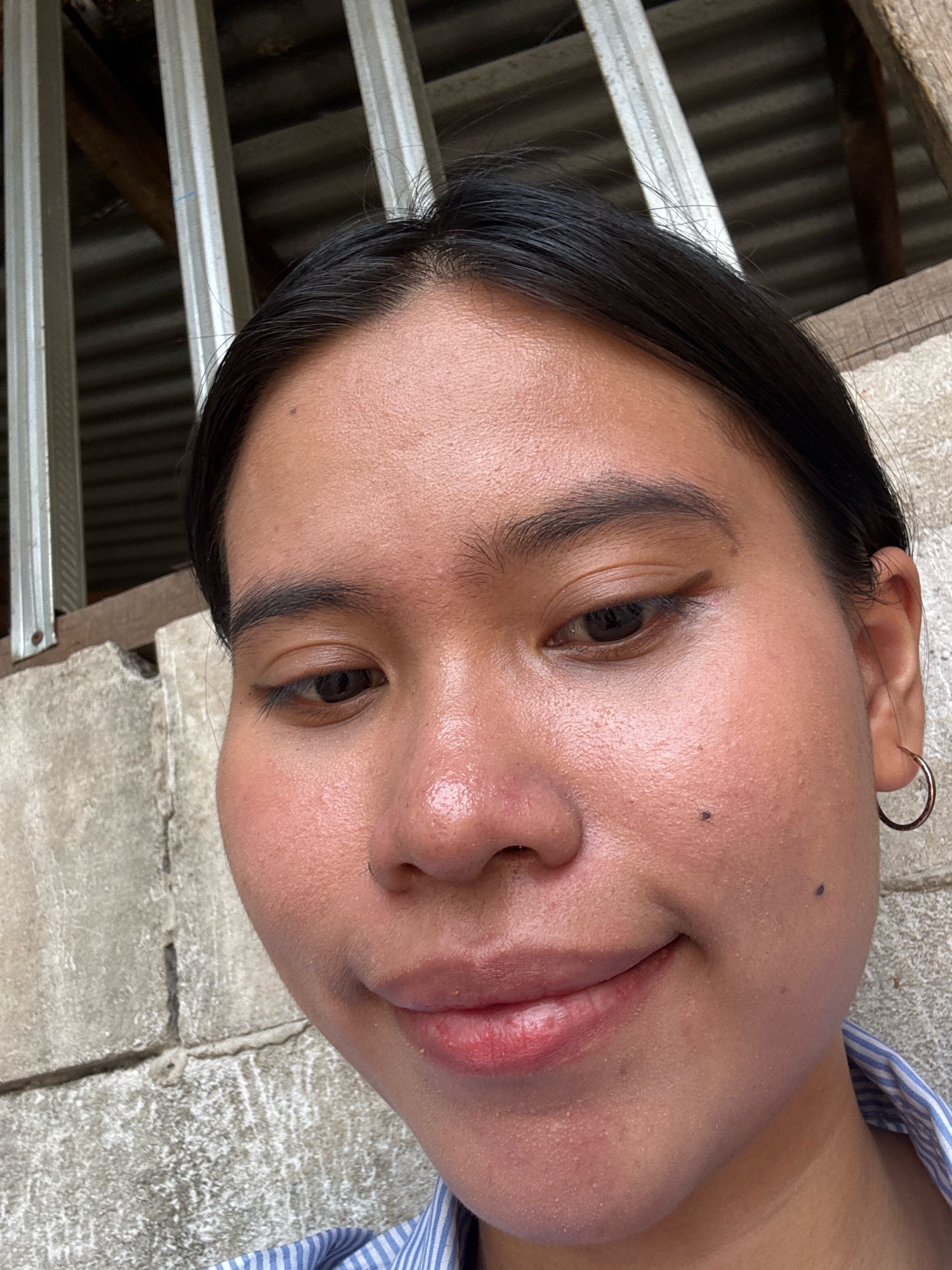 A close-up, eye-level, medium shot captures a young woman's face from the nose up, against a neutral background. She is looking down and slightly to her left. Her dark hair is pulled back, revealing her forehead and the sides of her face. Her skin appears to have a slight sheen, and there are a few small dark spots on her cheeks and forehead. She is wearing a striped shirt with a blue and white pattern, the collar of which is visible at the bottom of the frame. A thin hoop earring is visible in her right ear. The background consists of a textured, greyish-brown wall composed of rectangular blocks, and a corrugated metal roof structure with vertical metal bars visible behind her. The lighting suggests an outdoor setting, possibly during the day. The overall impression is a candid portrait with a focus on the subject's features.